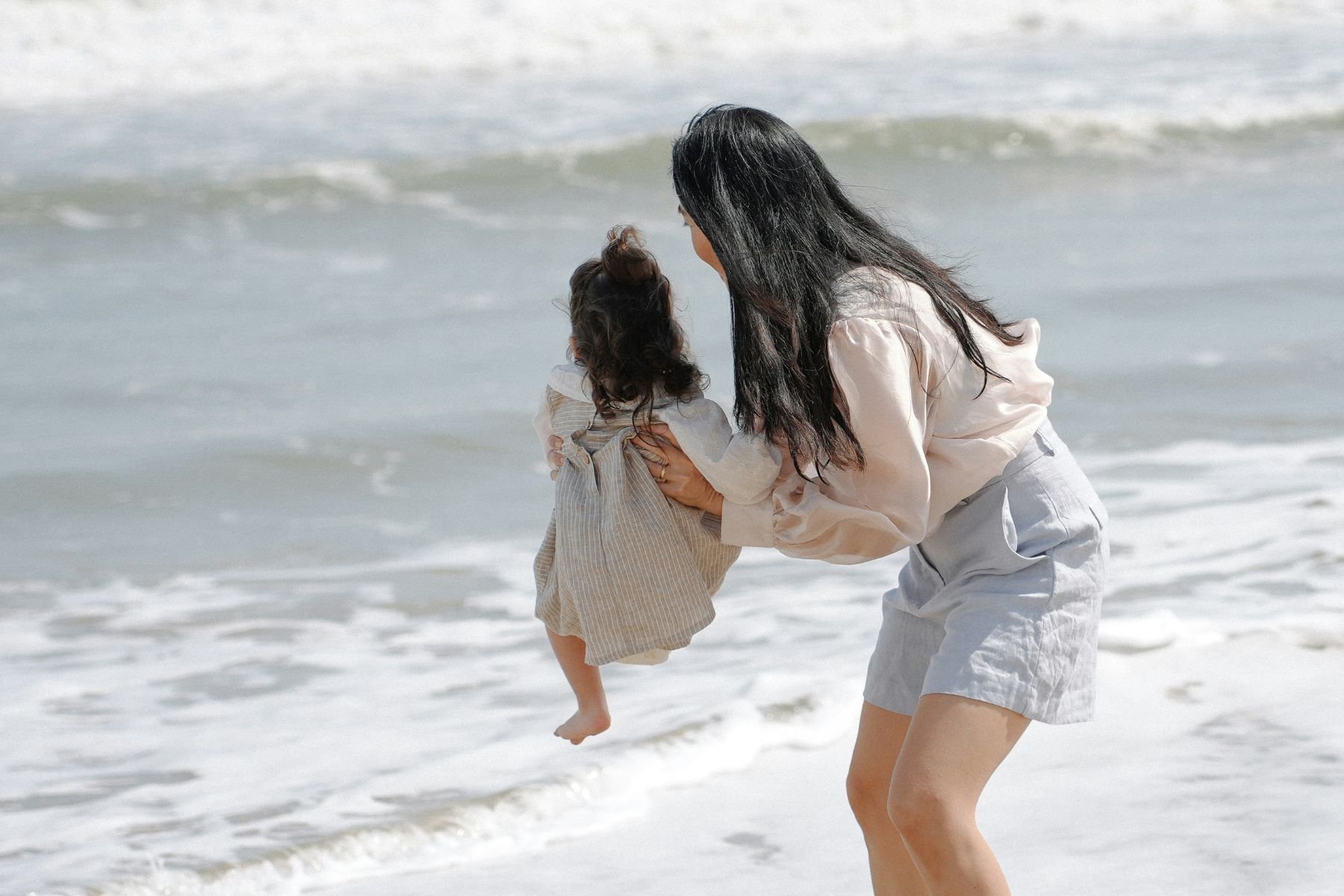 Woman holding a small child at the beach with ocean waves in the background.