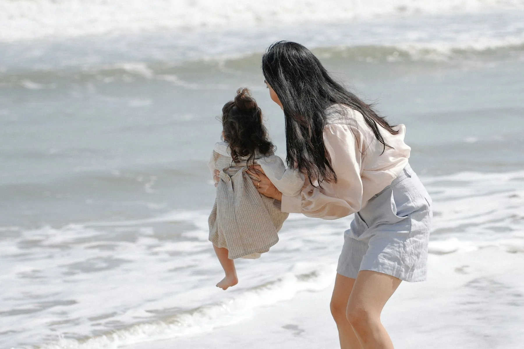 Woman holding a small child at the beach with ocean waves in the background.