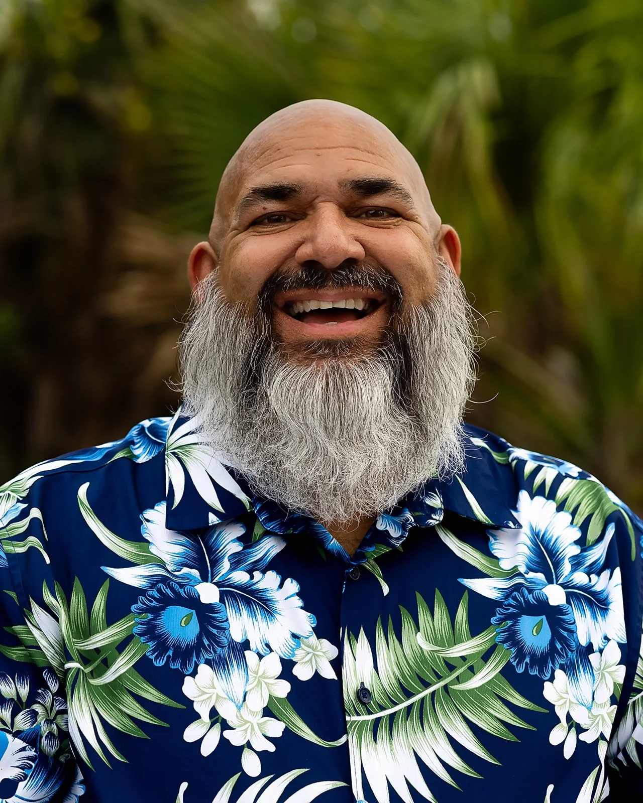 Stephen of Padre Island Baptist Church smiling with a long gray beard and bald head wearing a blue floral Hawaiian shirt, outdoors with green foliage in the background.