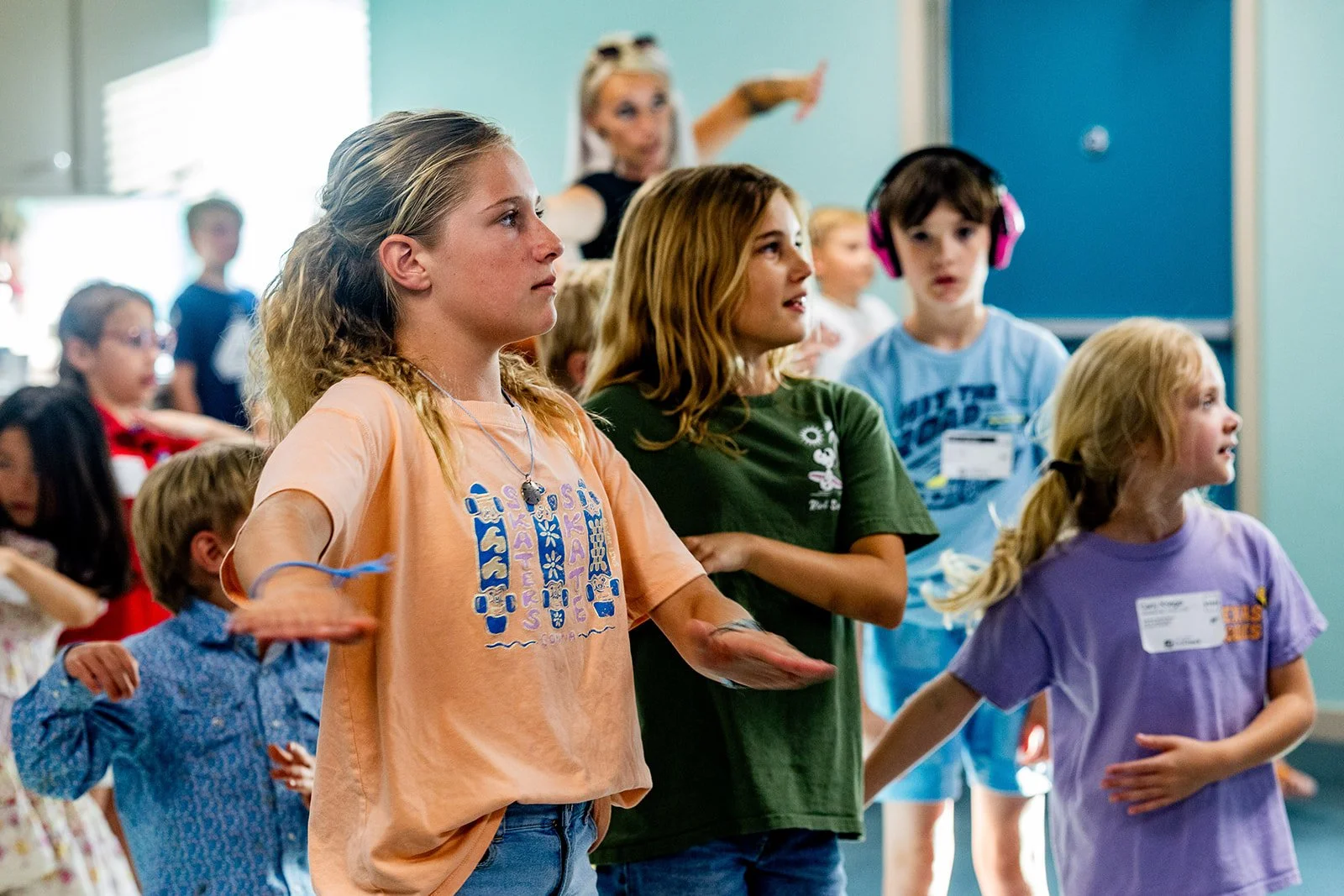A group of children and a woman participating in a communal activity indoors, with some children raising their hands and the woman guiding them.