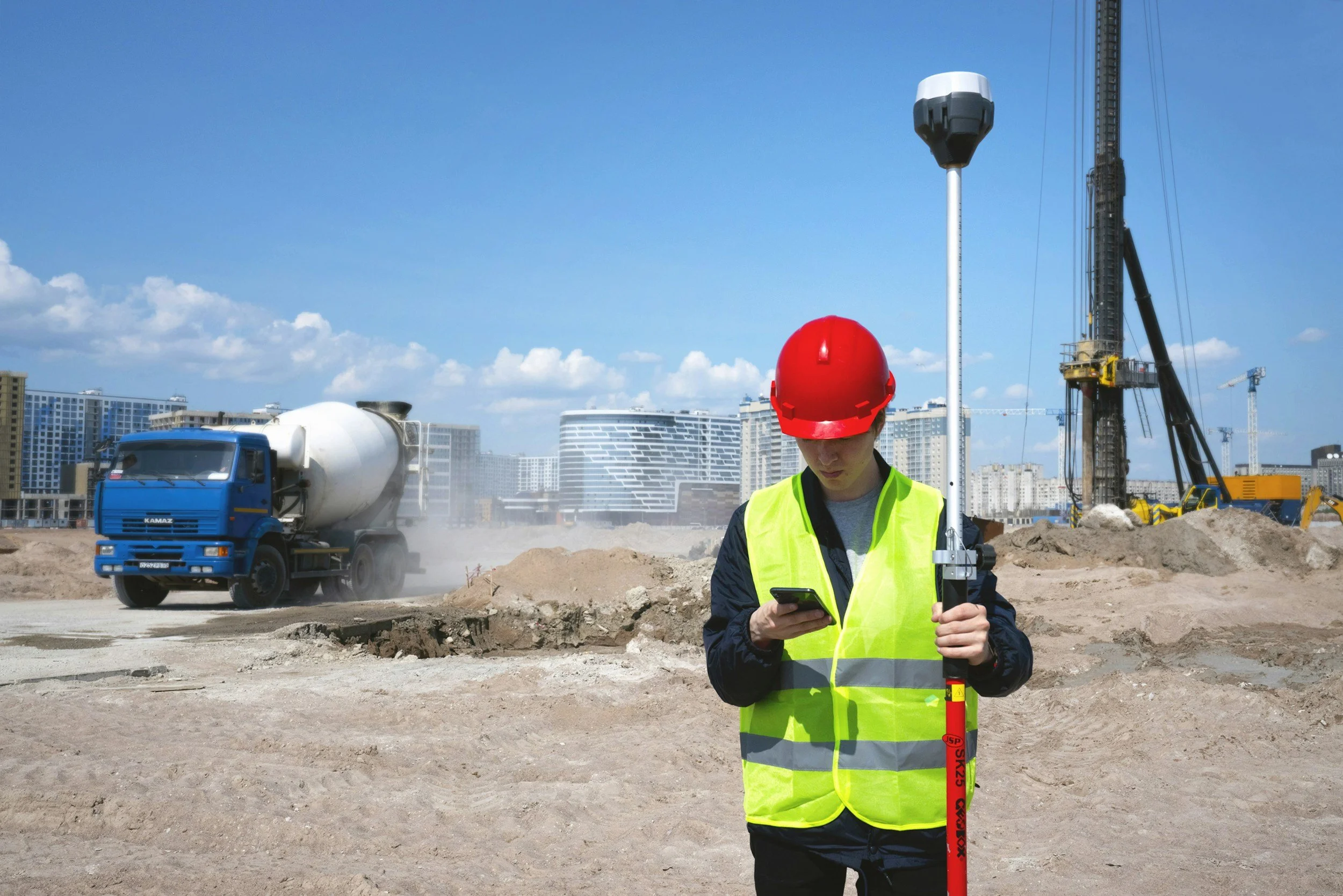 Construction worker in a yellow safety vest and red helmet using a surveying rod and phone at a construction site with a cement truck, heavy machinery, and city buildings in the background.