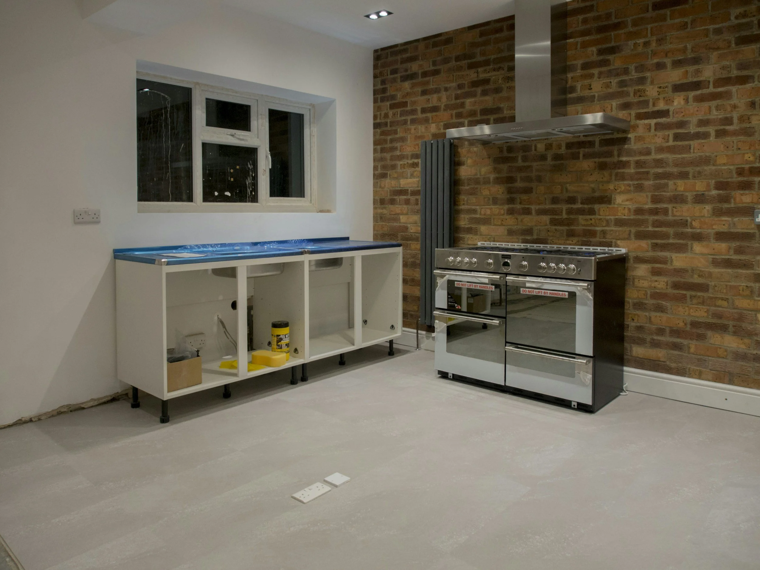 Unfinished kitchen with a brick accent wall, a window, and a stainless steel stove. There is an empty white cabinet base and some construction tools on the floor.