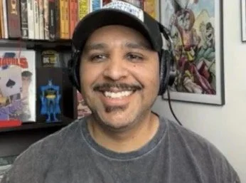 JUAN smiling, wearing headphones and a gray t-shirt, in front of a bookshelf with comic book covers and posters.