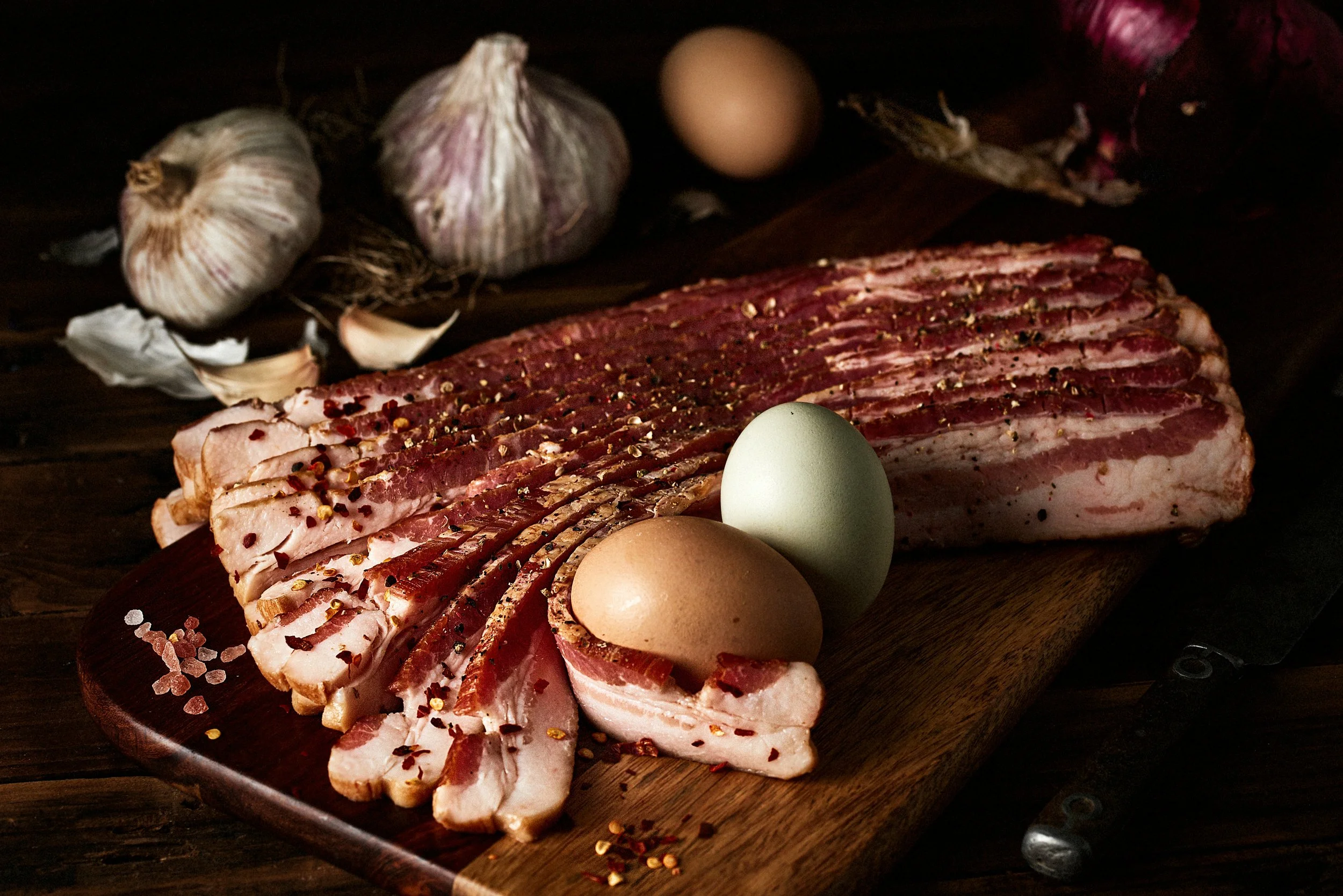 Sliced cured pork belly on a wooden cutting board, accompanied by two eggs and garlic bulbs in the background.
