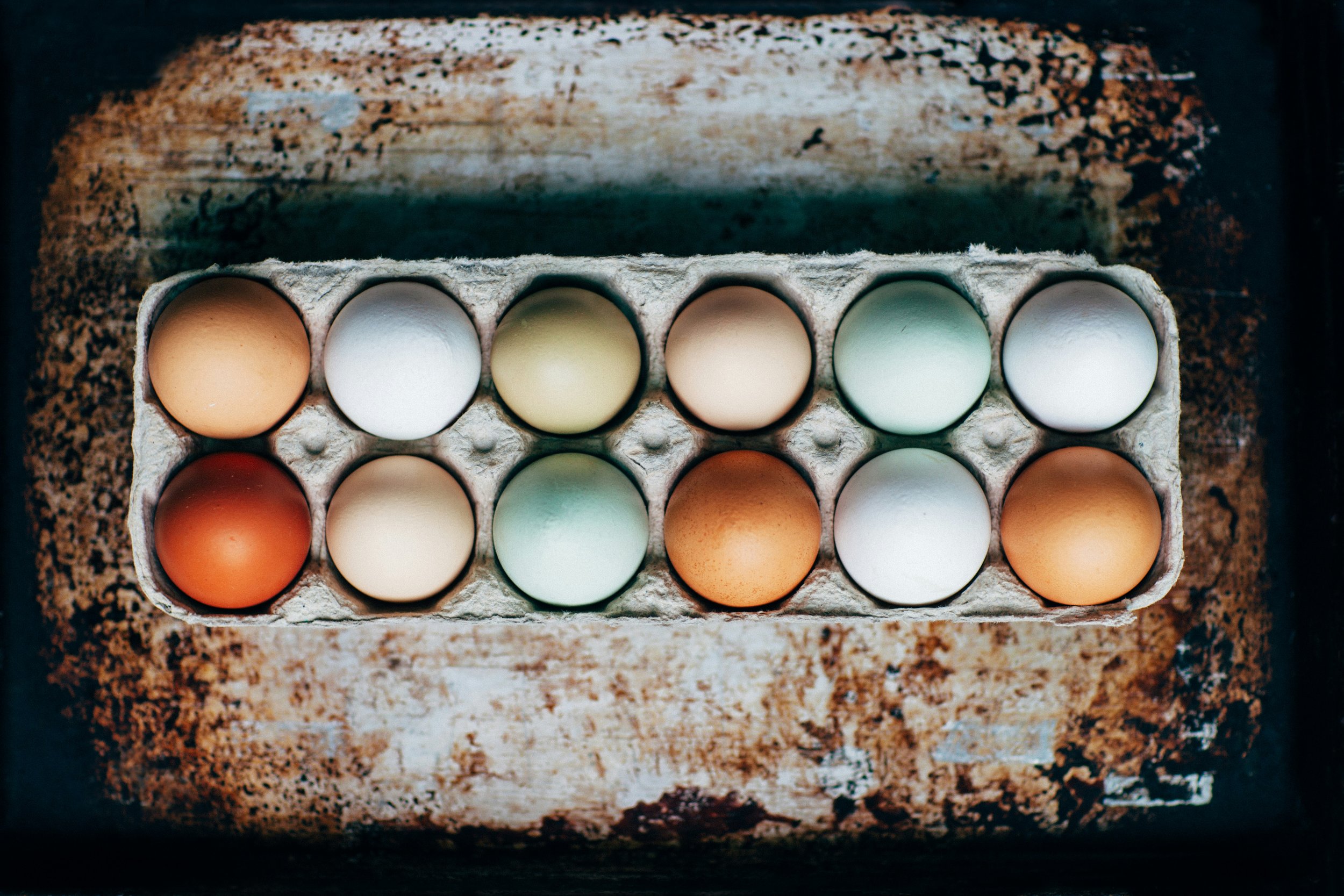 A carton of twelve eggs with various colors, placed on a rusty metal surface.