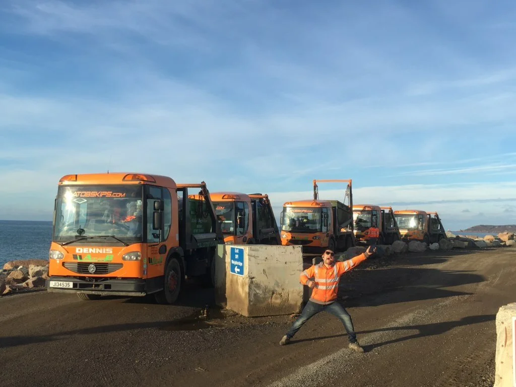 A man in an orange safety jacket posing excitedly in front of a row of five orange street sweepers near the coastline with rocks and water in the background.
