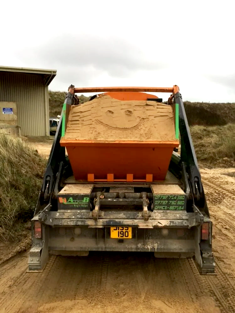 A construction or demolition site showing a vehicle with a large wooden crate on its back, crafted to resemble a smiling face with a wide grin. The vehicle is parked on dirt ground near a building and grassy area, under cloudy skies.