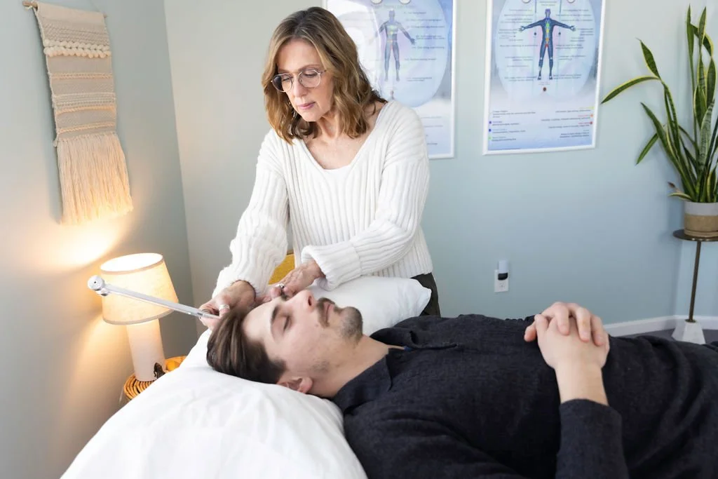A professional sound healer woman appears to be performing a therapy or medical procedure on a man lying in a bed, in a room with medical charts.