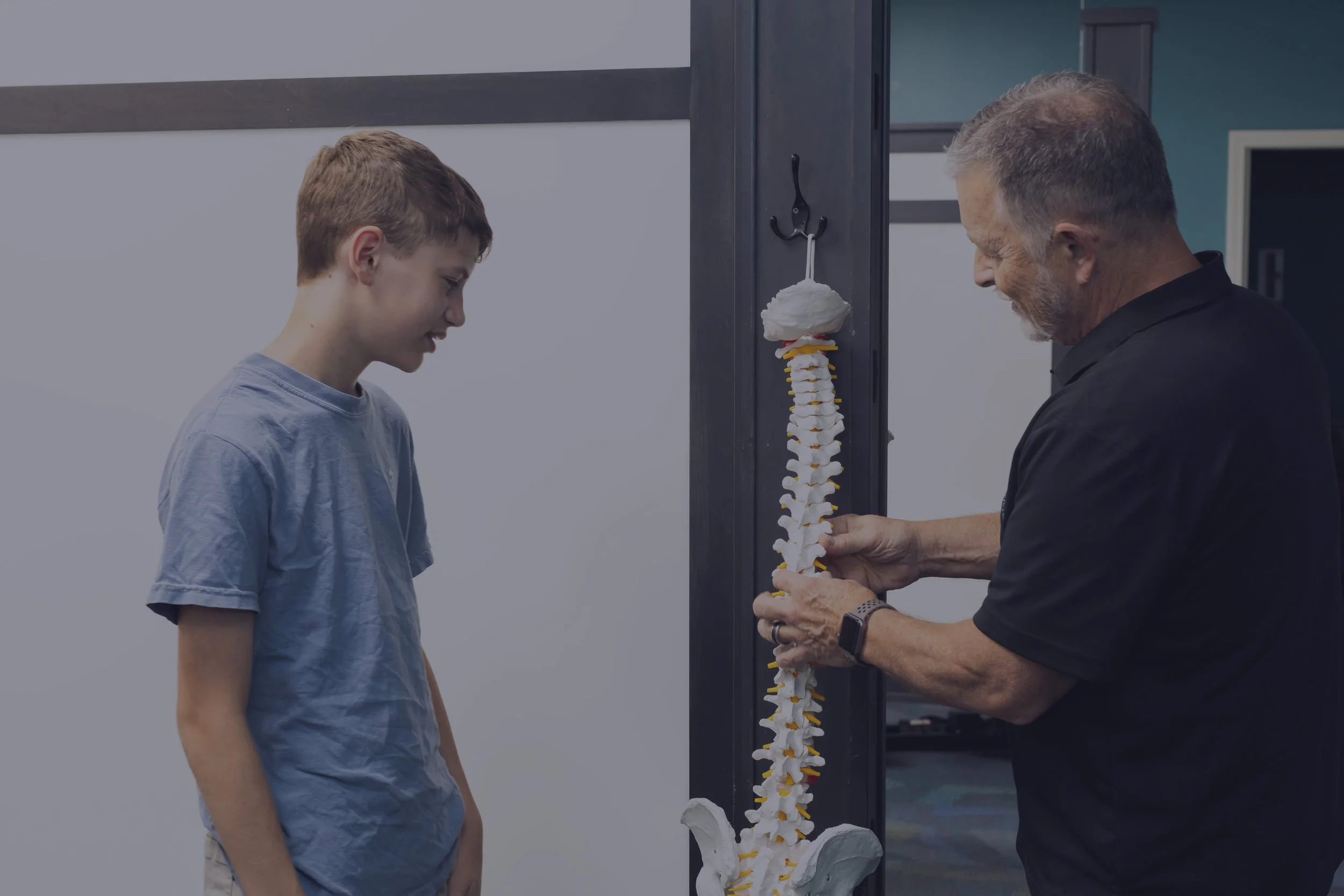 A young boy and an older man looking at a model of a human spine in a clinical setting.
