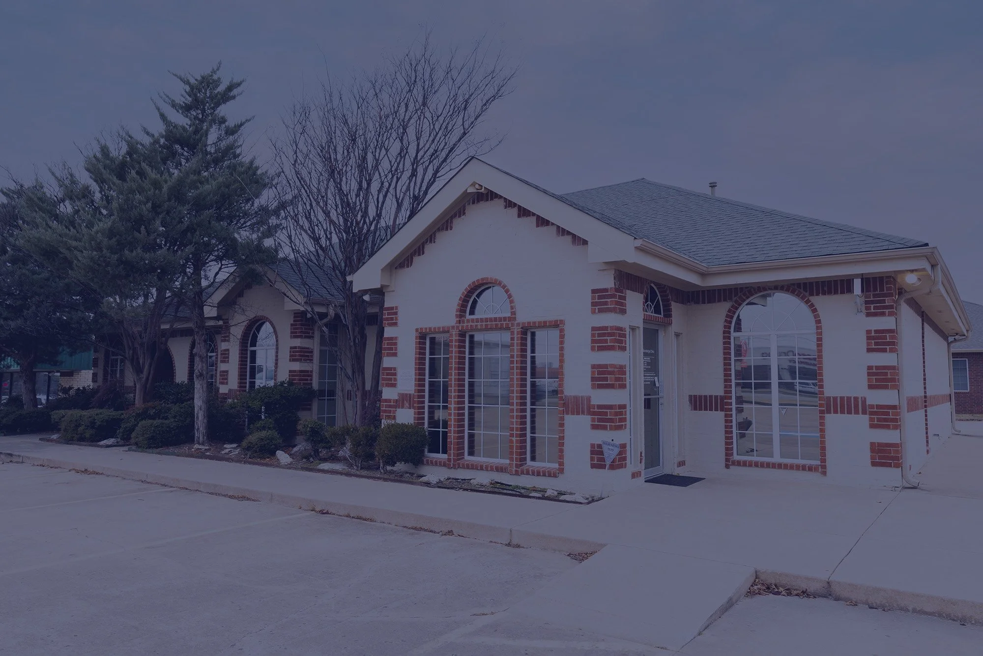 A single-story commercial building with white walls accented by red brick trim, large arched windows, and surrounded by a parking lot with a sidewalk and landscaping with trees and bushes.