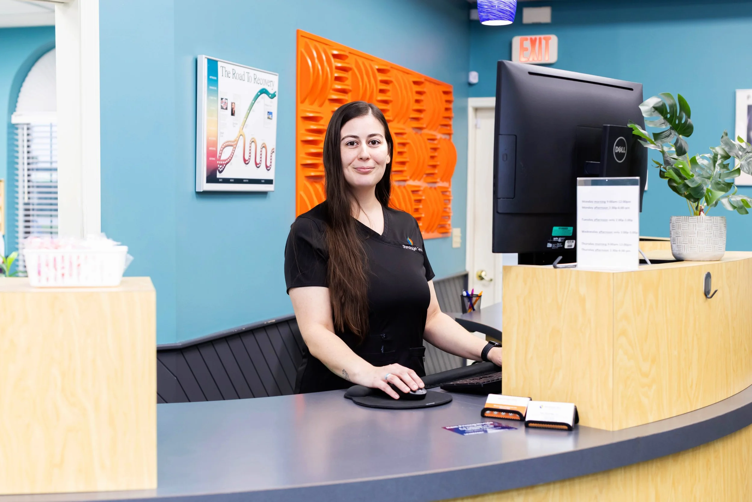 A woman with long dark hair standing at a reception desk, smiling, with a computer, potted plant, and informational sign on the desk. The background includes a blue wall, an orange decorative panel, and a framed chart.