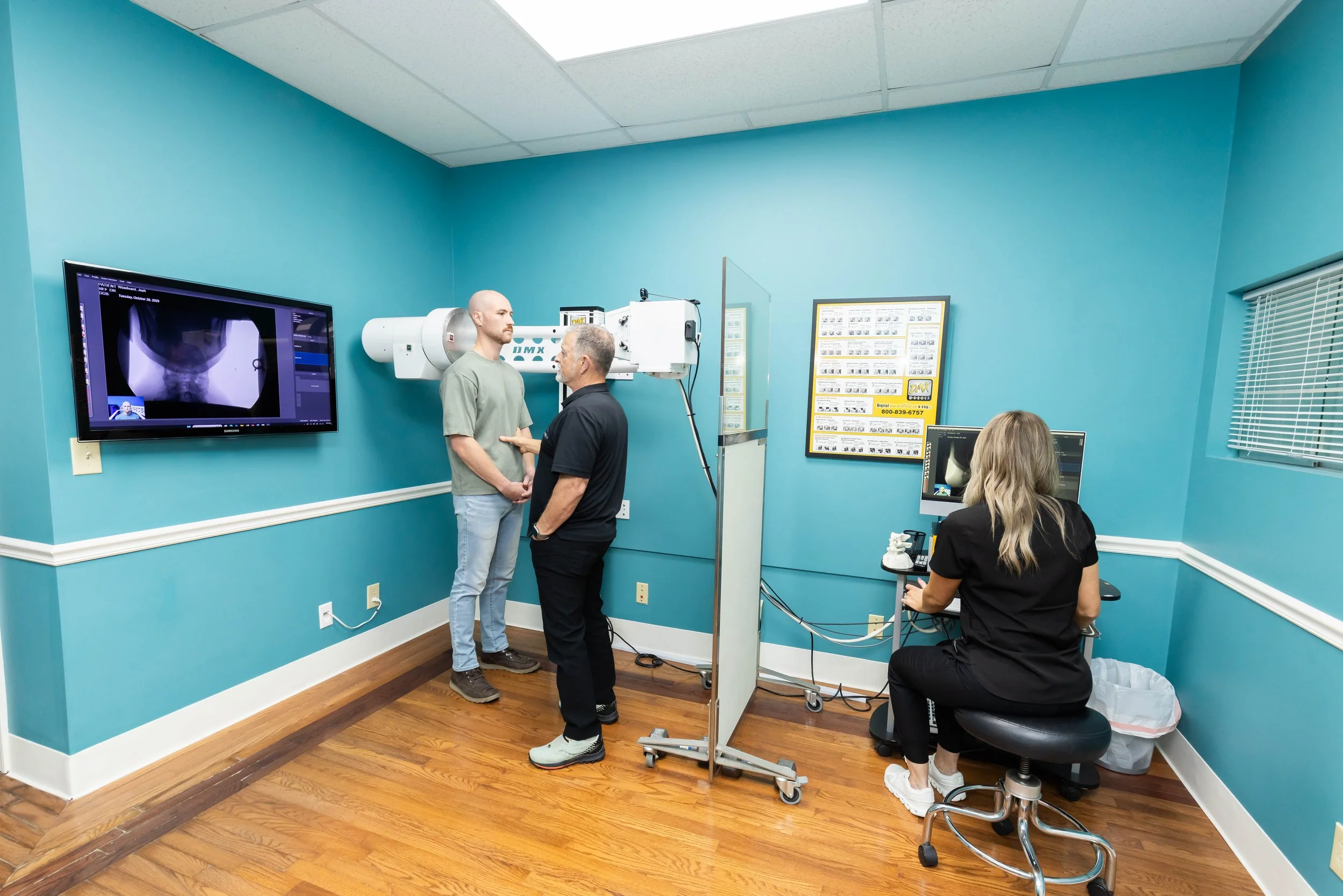 A medical imaging room with a patient standing with a doctor while a technician operates a machine, and a technician monitors imaging on a computer.