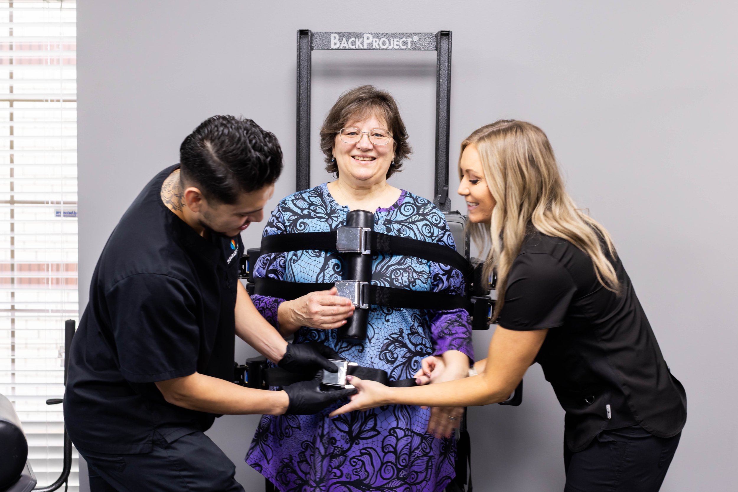 A woman in a harness being strapped into a back brace by two healthcare workers in a clinical setting.