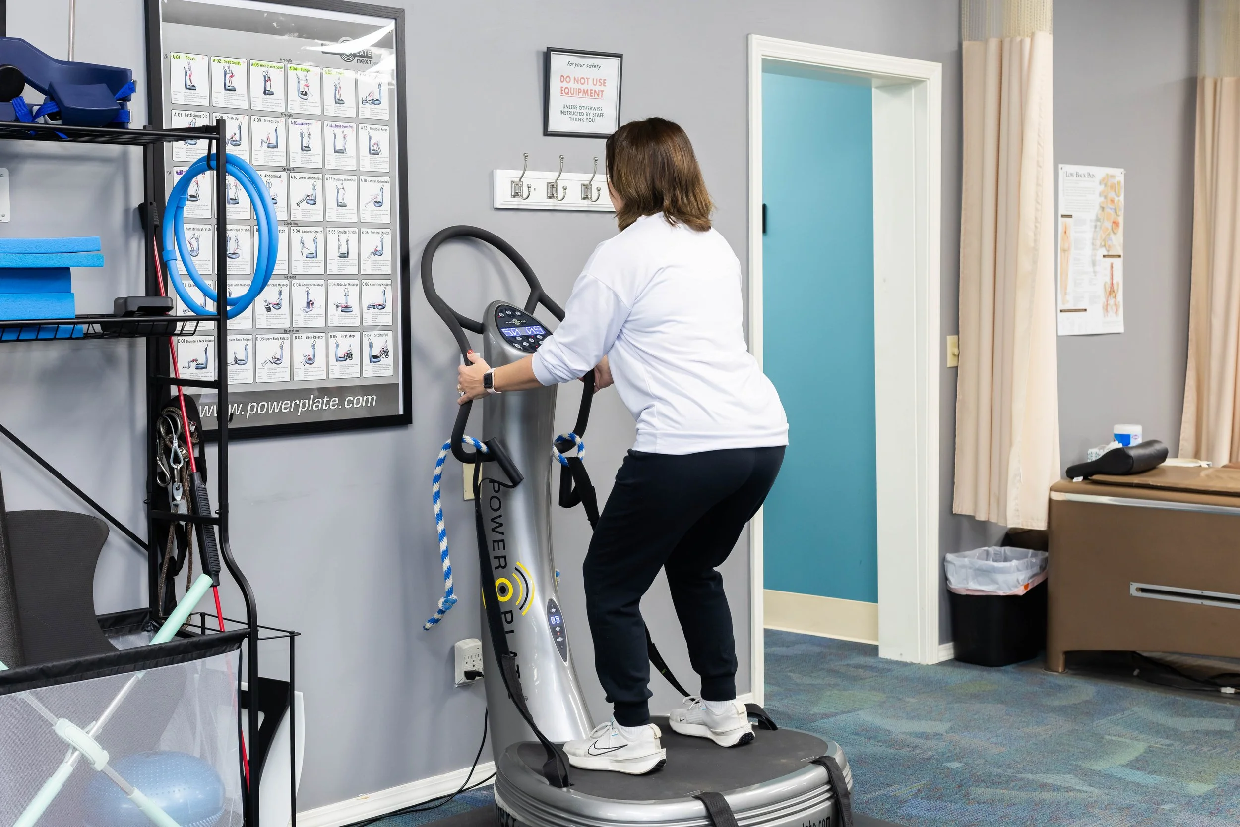 A woman in a white shirt and black pants standing on a vibration plate exercise machine in a physical therapy or gym room.