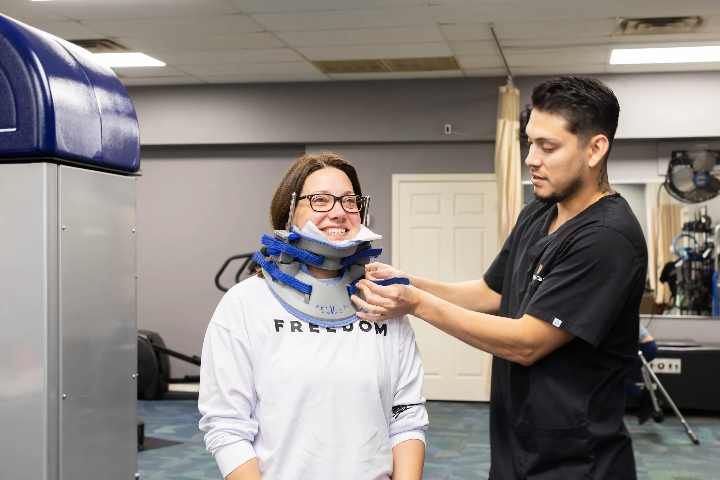 A woman in a white shirt with the word 'FREEDOM' on it is smiling while a healthcare worker helps her put on a neck brace in a clinic or physical therapy setting.