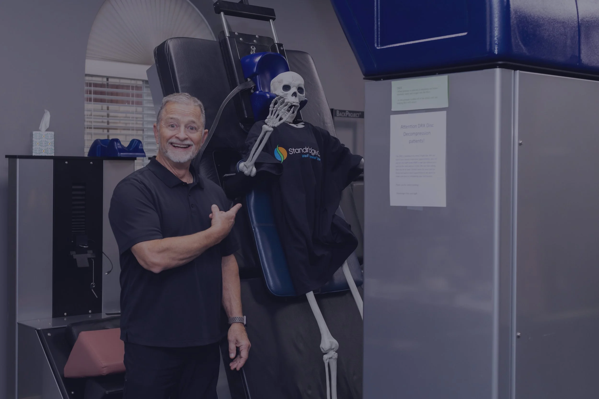 A man smiling and pointing at a skeleton mannequin sitting in a medical recliner chair with a black t-shirt and a skull mask, in a clinic room.
