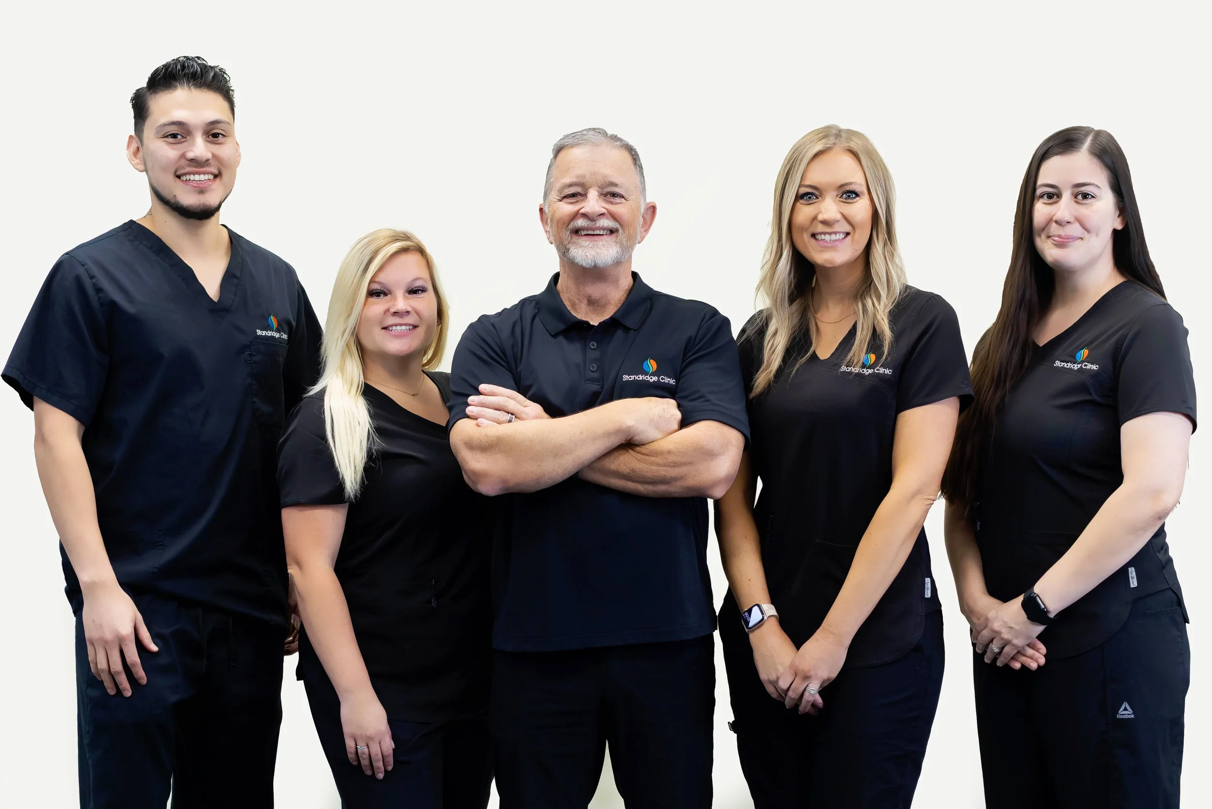 A group of five healthcare professionals in black uniforms with a logo, standing together and smiling against a white background.