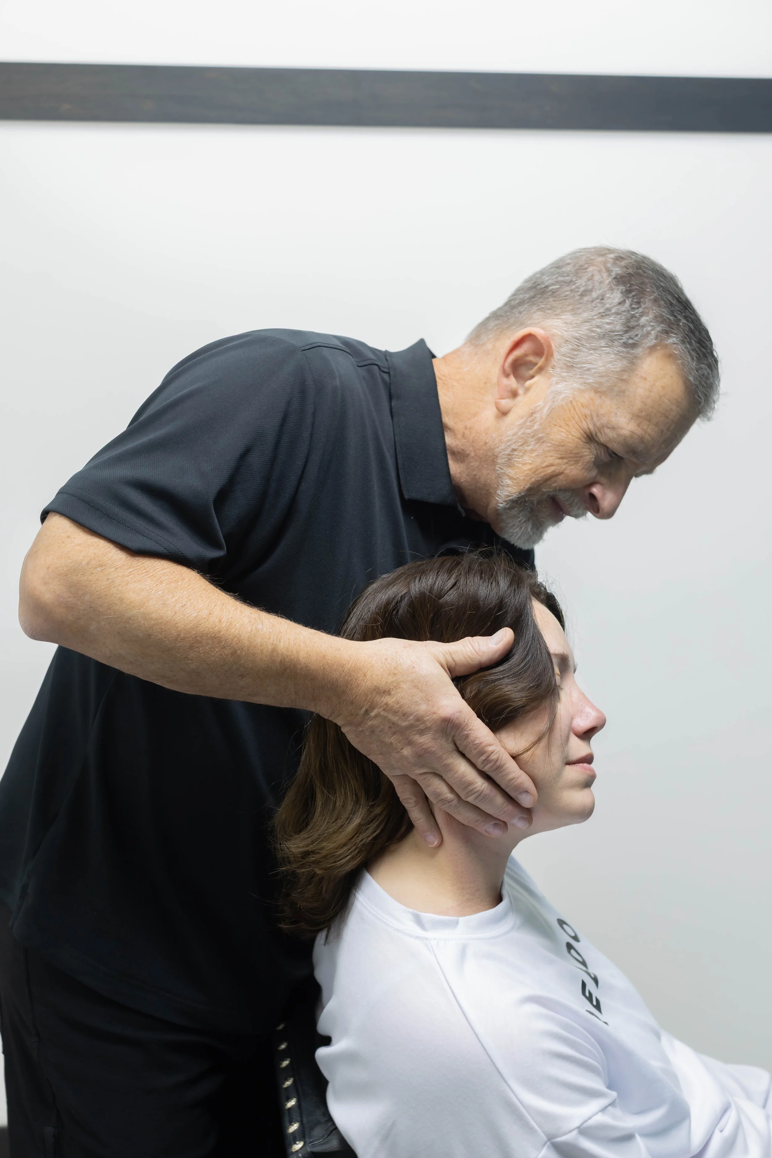 An older man with gray hair and beard gently holding a young woman's head, providing chiropractic or massage therapy in a clinical setting.