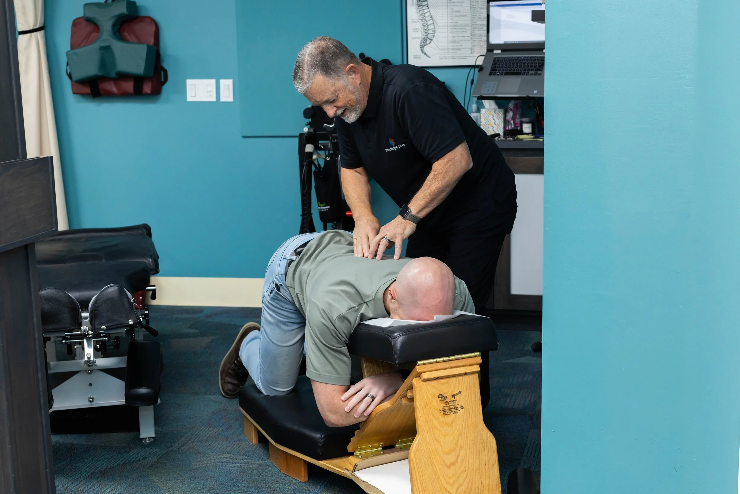 A chiropractor adjusting a patient's back while the patient lies face down on a chiropractic table in a clinic.