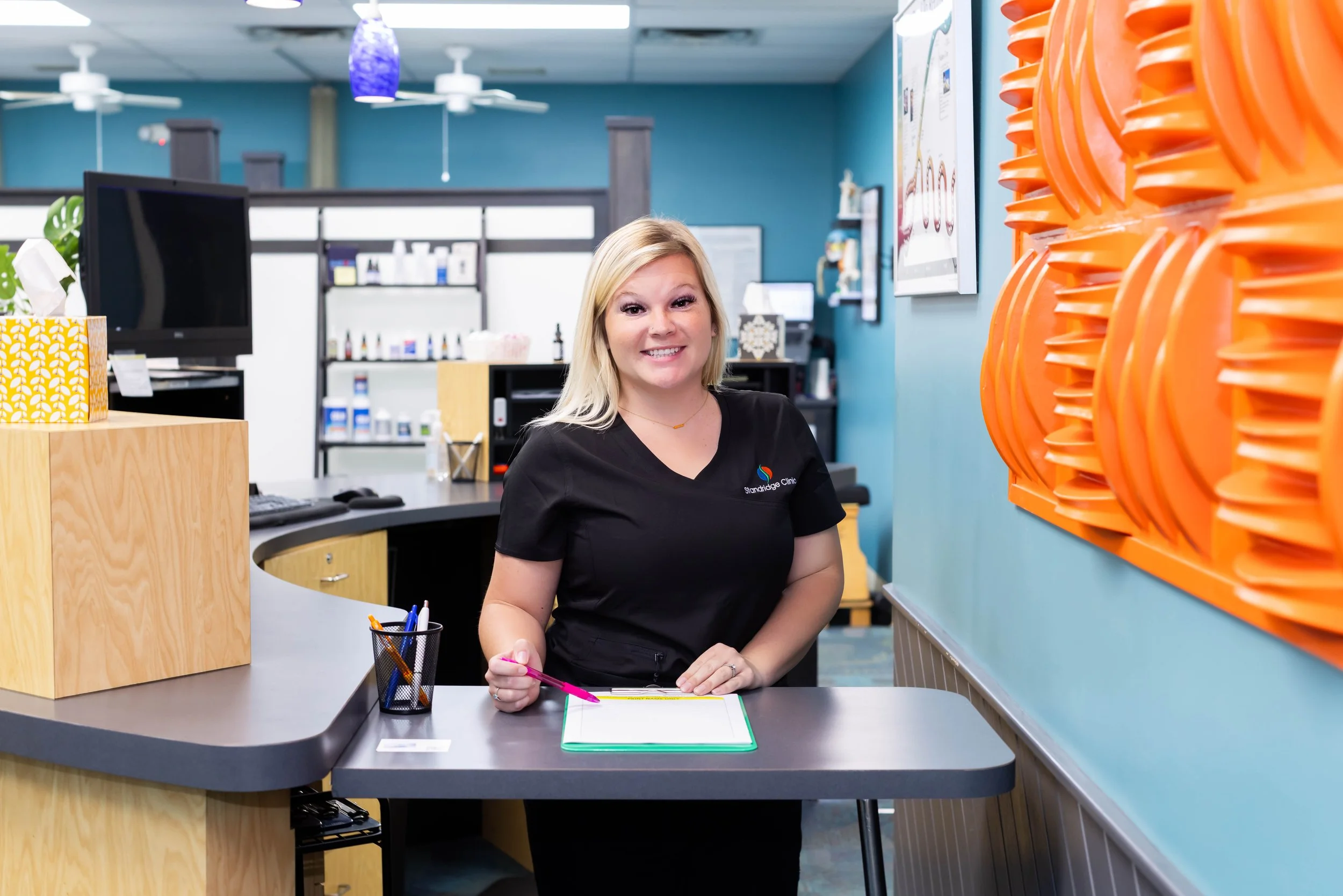 A smiling woman with blonde hair wearing a black medical uniform sitting at a reception desk with a clipboard and pink pen in a medical office or clinic.