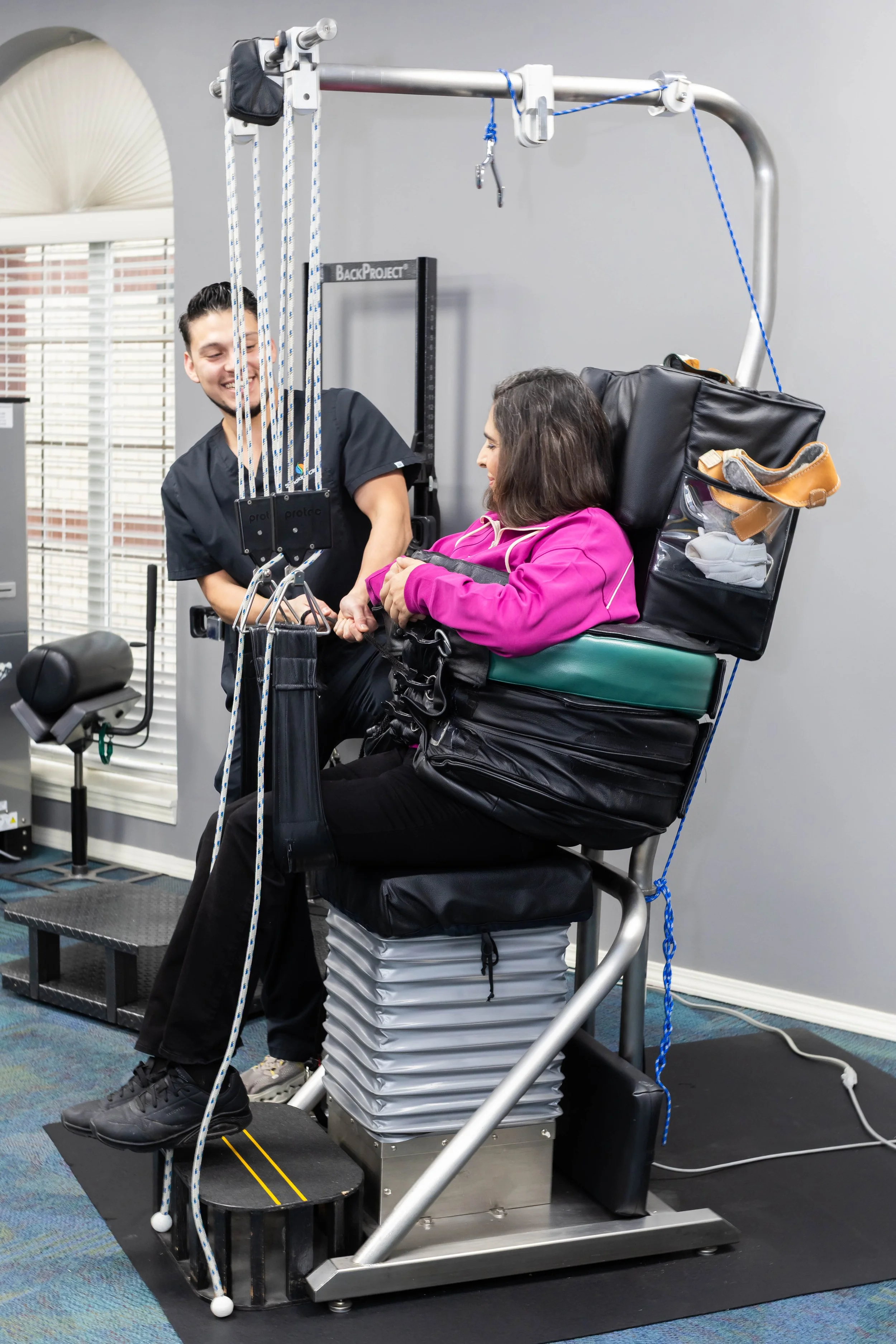 A woman seated in a specialized medical or rehabilitation device, being assisted by a healthcare professional, in a clinical setting with window blinds and exercise equipment visible.