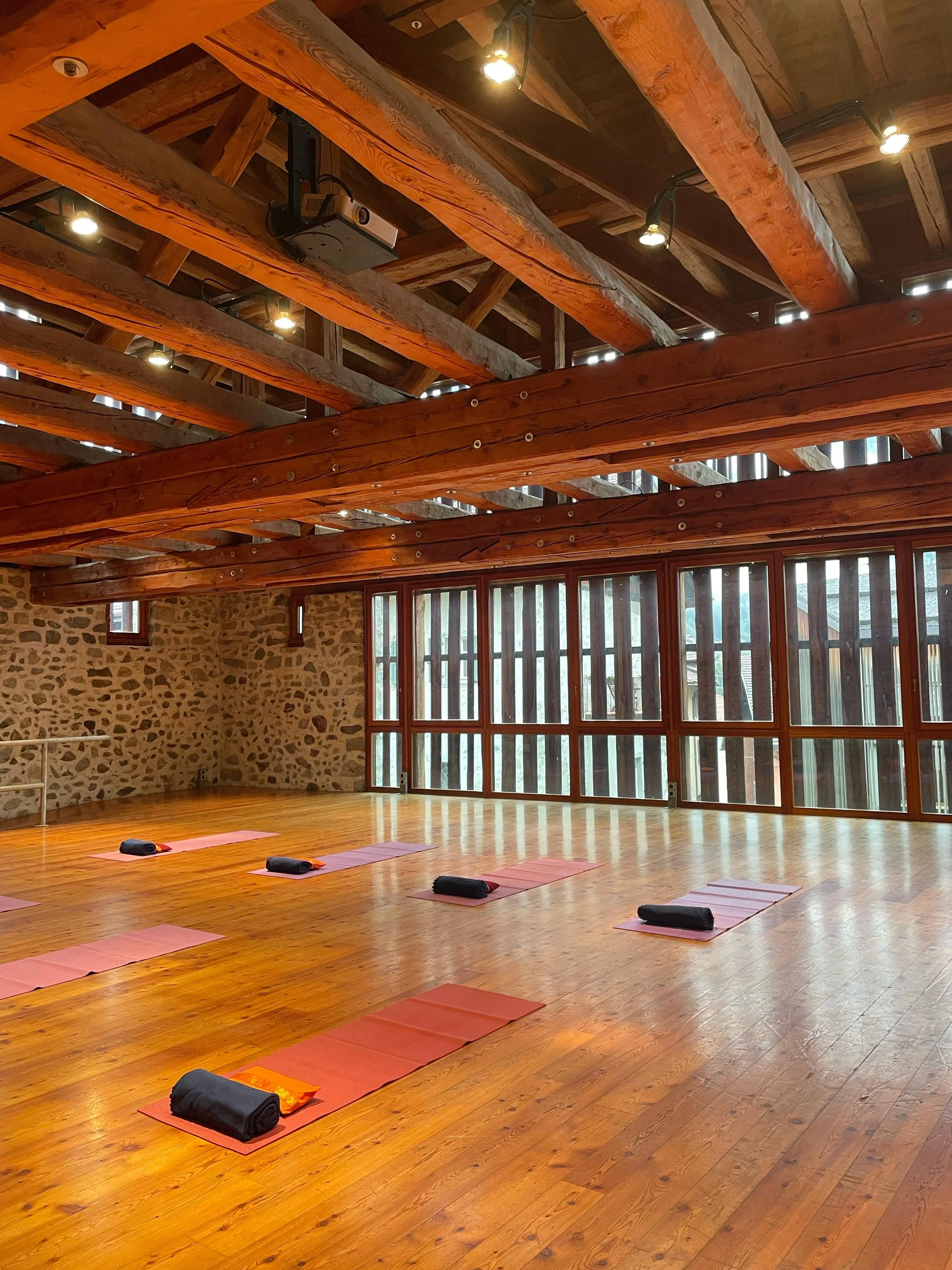 Sala per yoga o meditazione con tappetini arancioni, copertine nere e blocchi sopra di essi, grande finestra con pannelli di vetro, pareti in pietra e soffitto in legno con travi di supporto.