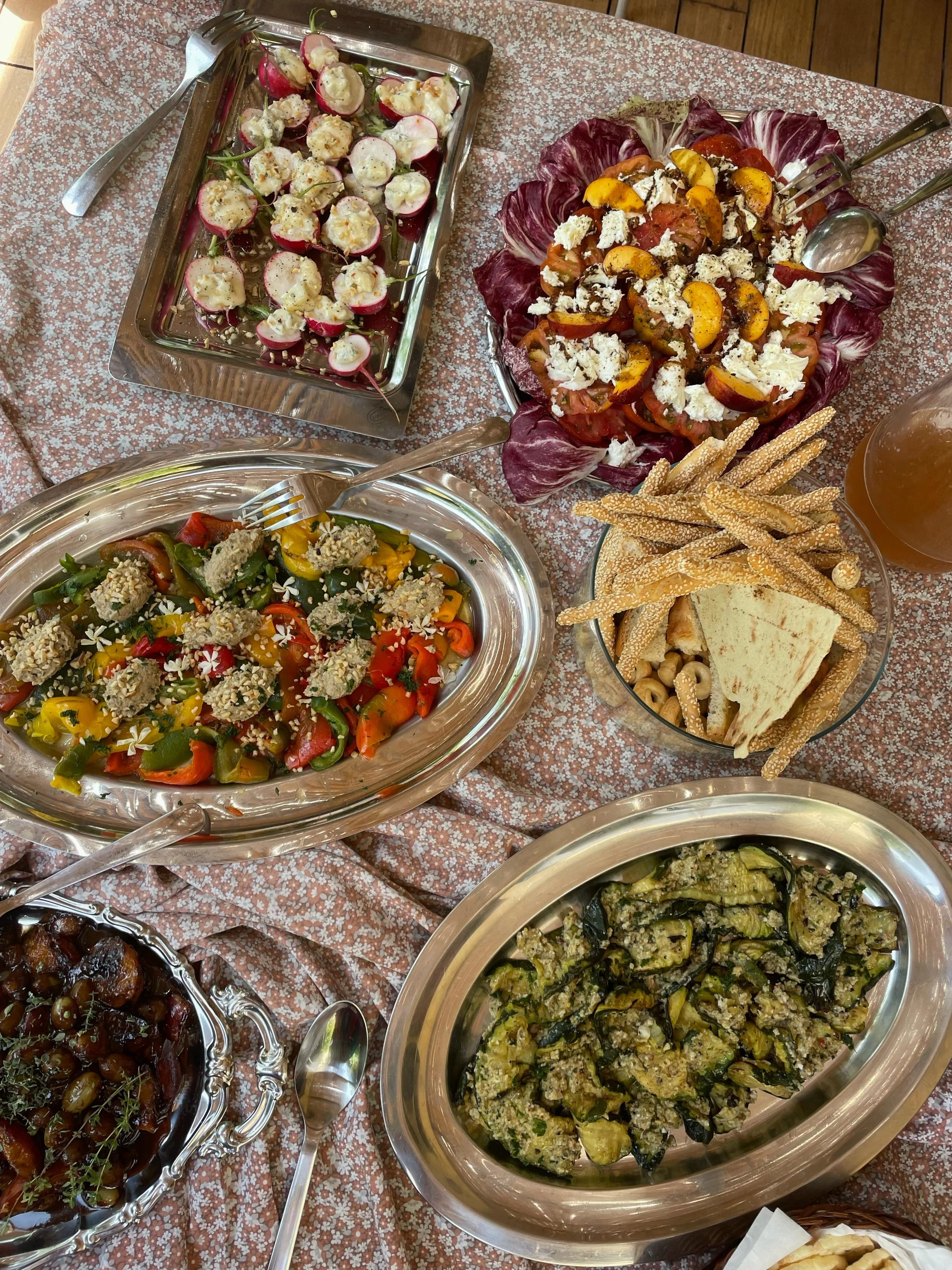 Varie piatti di insalate e verdure servite in ciotole e piatti di metallo, con utensili. Cibi come insalate di pomodori, melanzane e castagne, e patatine di sesamo, su una tovaglia decorata.
