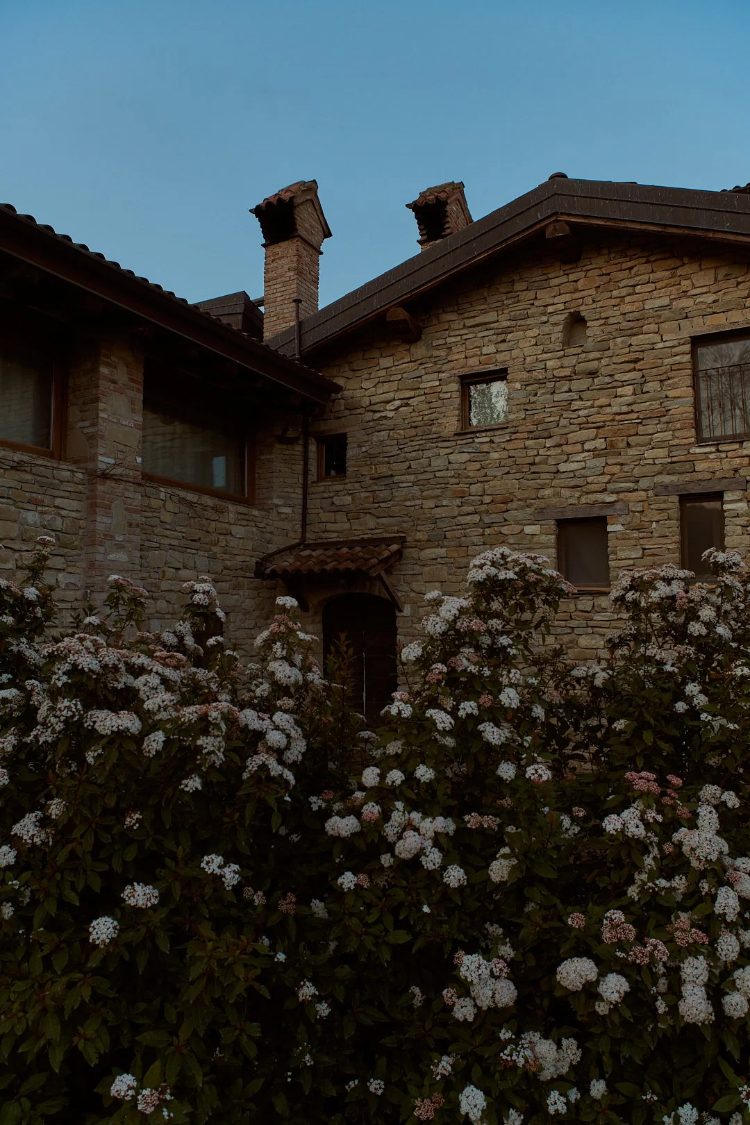 Casa in pietra con finestre e camino, circondata da fiori di color bianco e rosa, sotto un cielo blu