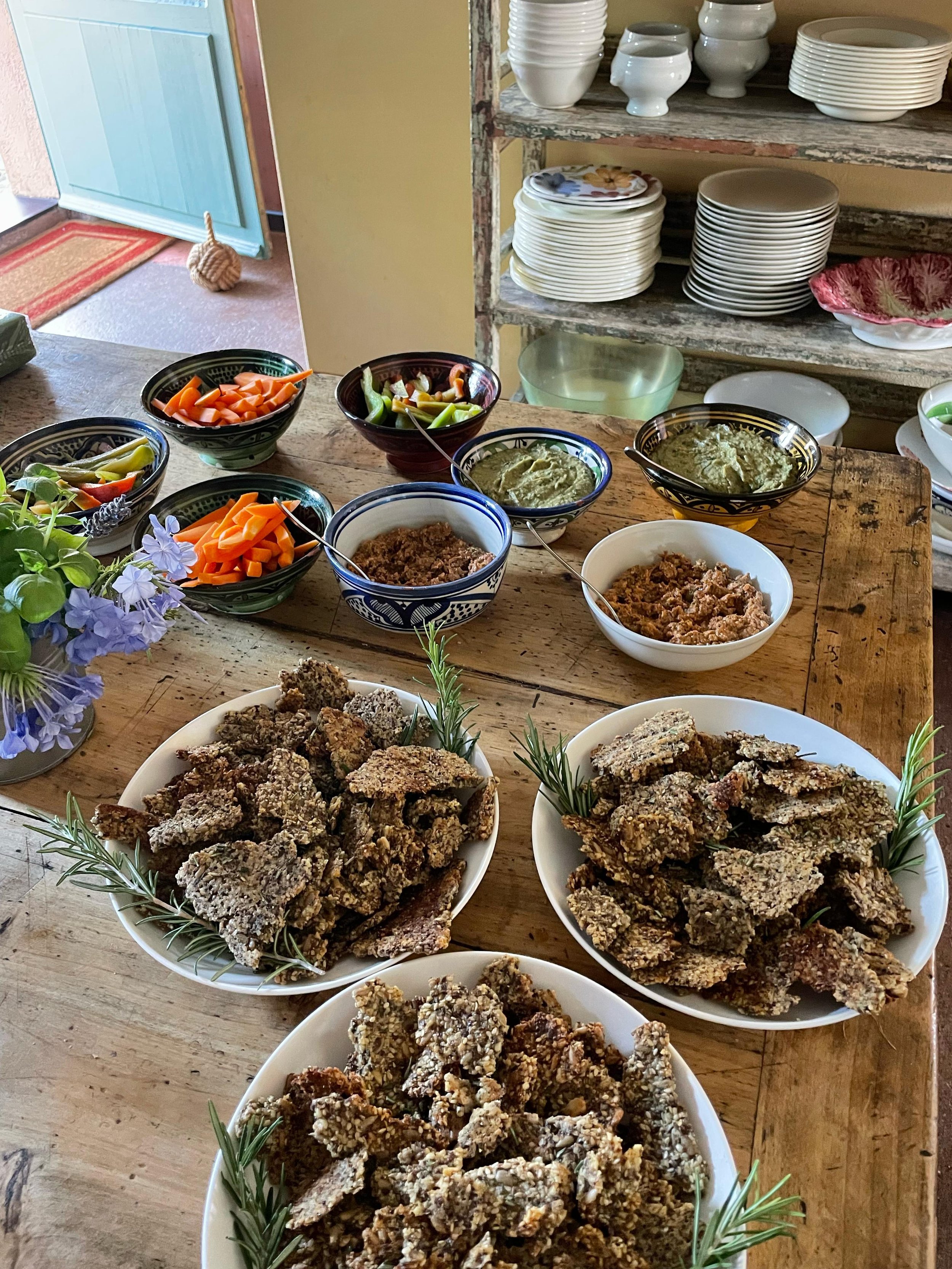 Una tavola imbandita con diverse ciotole di vario cibo, tra cui salse, verdure e snack di pane o grissini, con piante decorative ai lati. In background, armadi e scaffali con piatti e bicchieri.