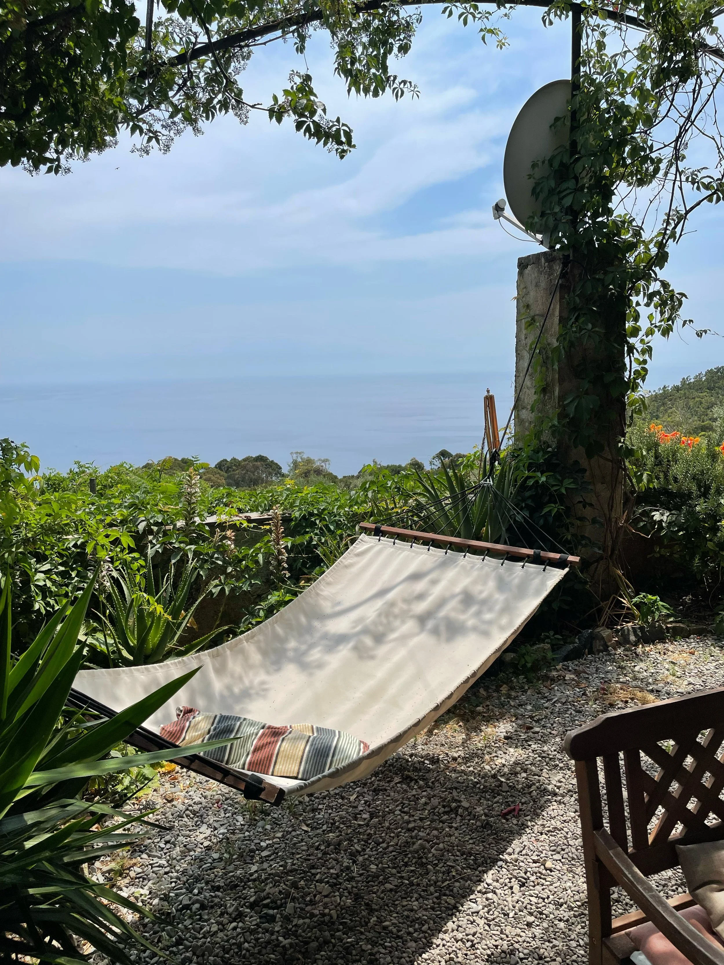 Un'area all'aperto con una sedia a sdraio con coperta a righe, piante verdi, un albero con rami e foglie, un muretto e uno spazio con ciottoli, con vista sul mare in lontananza