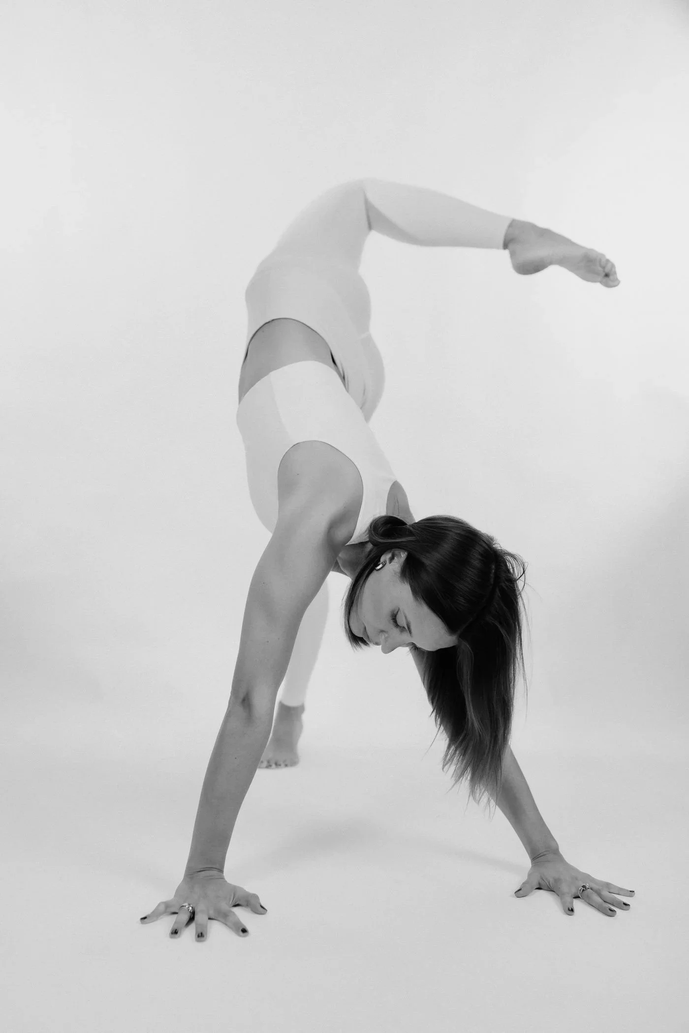 Donna che fa una posizione di yoga a testa in giù, con le mani e i piedi poggiati a terra, i capelli sciolti verso il basso, in un ambiente bianco.