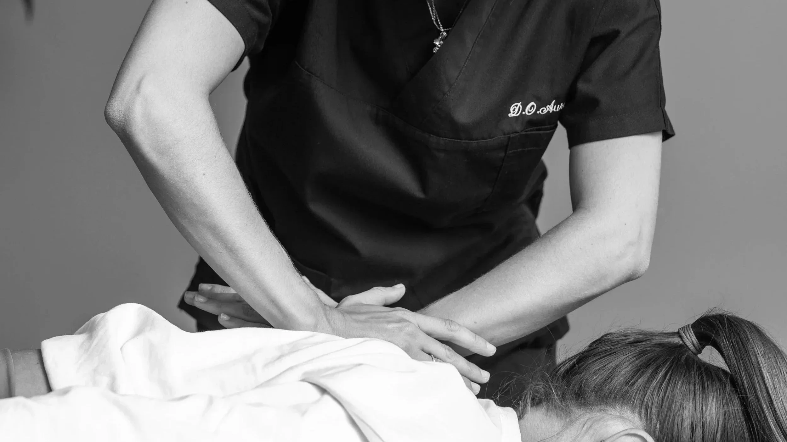 Un'operatrice sanitaria con camice nero che musica un braccio di una paziente durante una terapia o una visita medica.