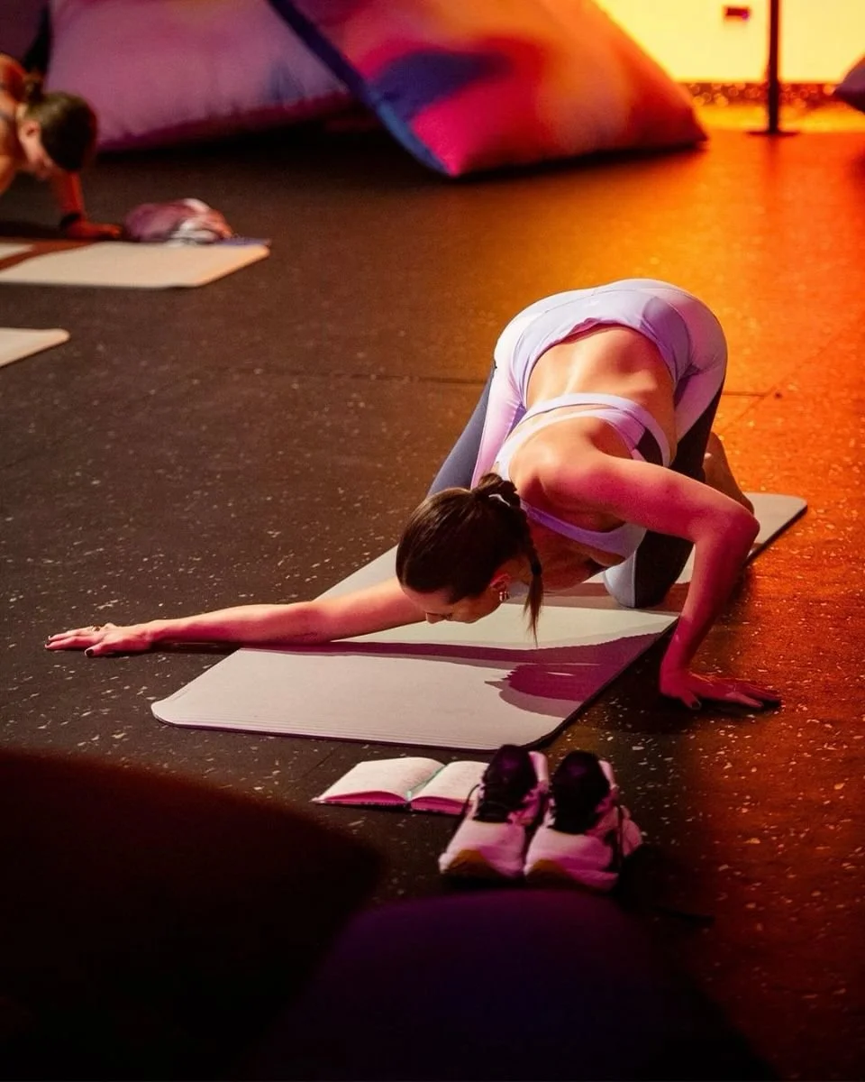 Una donna fa yoga in un'illuminazione calda, con una donna più in fondo che si allena vicino a un cuscino rosso e un libro aperto davanti a lei.