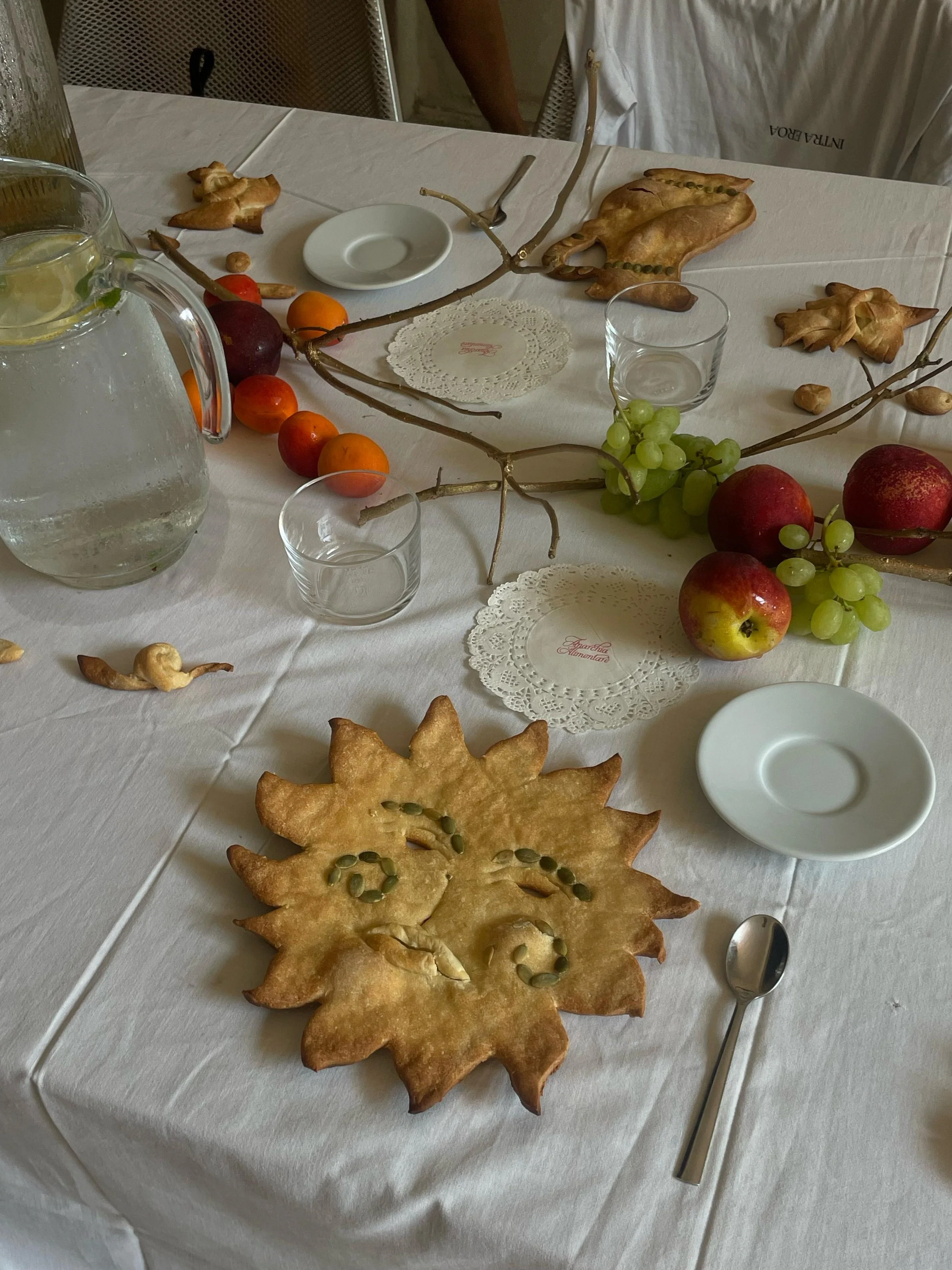 Tavolo imbandito con pane a forma di fiore decorato con semi di zucca, frutta come uva, mele e albicocche, acqua con limone, piatti e bicchieri trasparenti, cucchiaino e tovaglioli di carta.