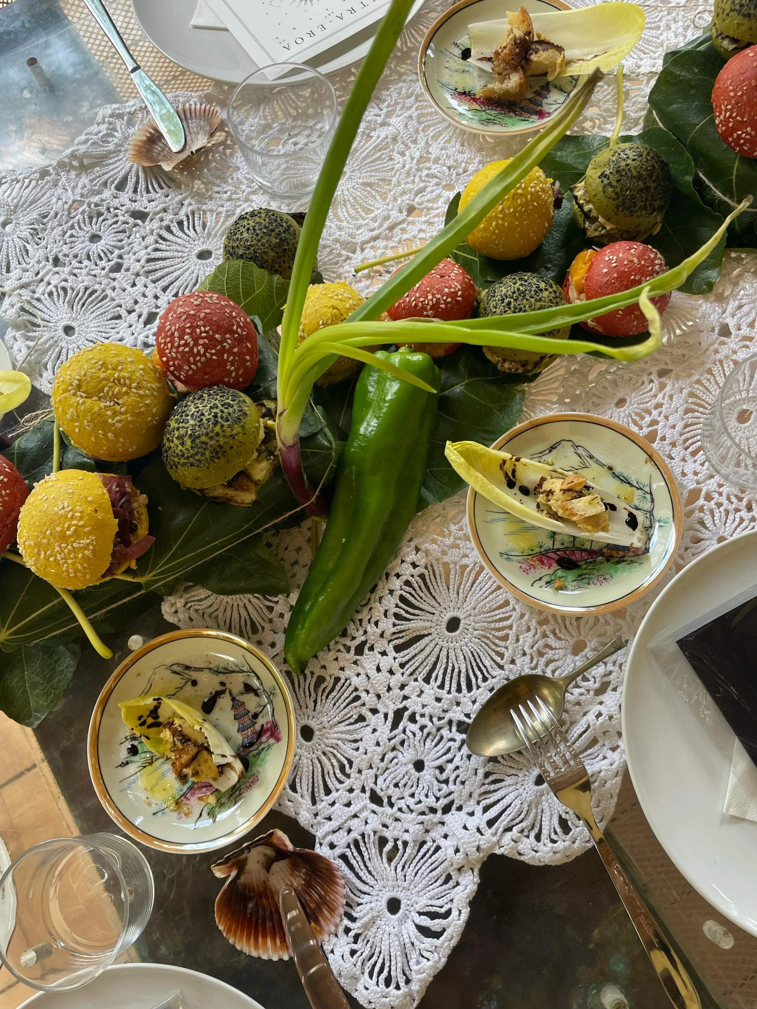 Una tavola apparecchiata con circa dieci ciotole e piatti decorativi, alcune contenenti resti di cibo, e molte conchiglie marine e uova di capodoglio, decorazioni naturali e pianta con foglie verdi, su tovagliolo di pizzo bianco.