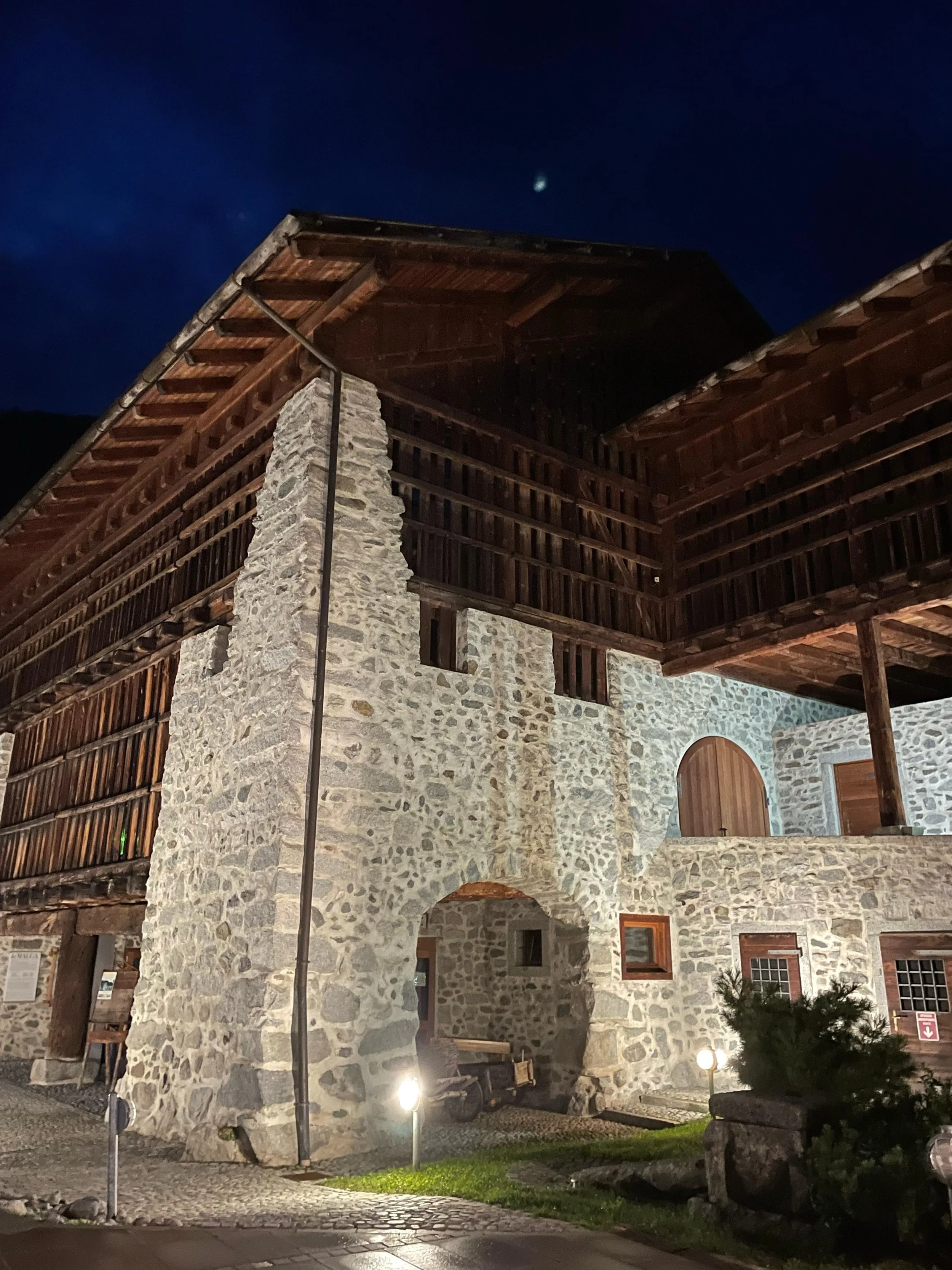 Edificio in pietra con balcone in legno sotto un cielo notturno, alcune luci illuminano l'area esterna. C'è un albero e un lampione. Nella notte si intravede la luna nel cielo.