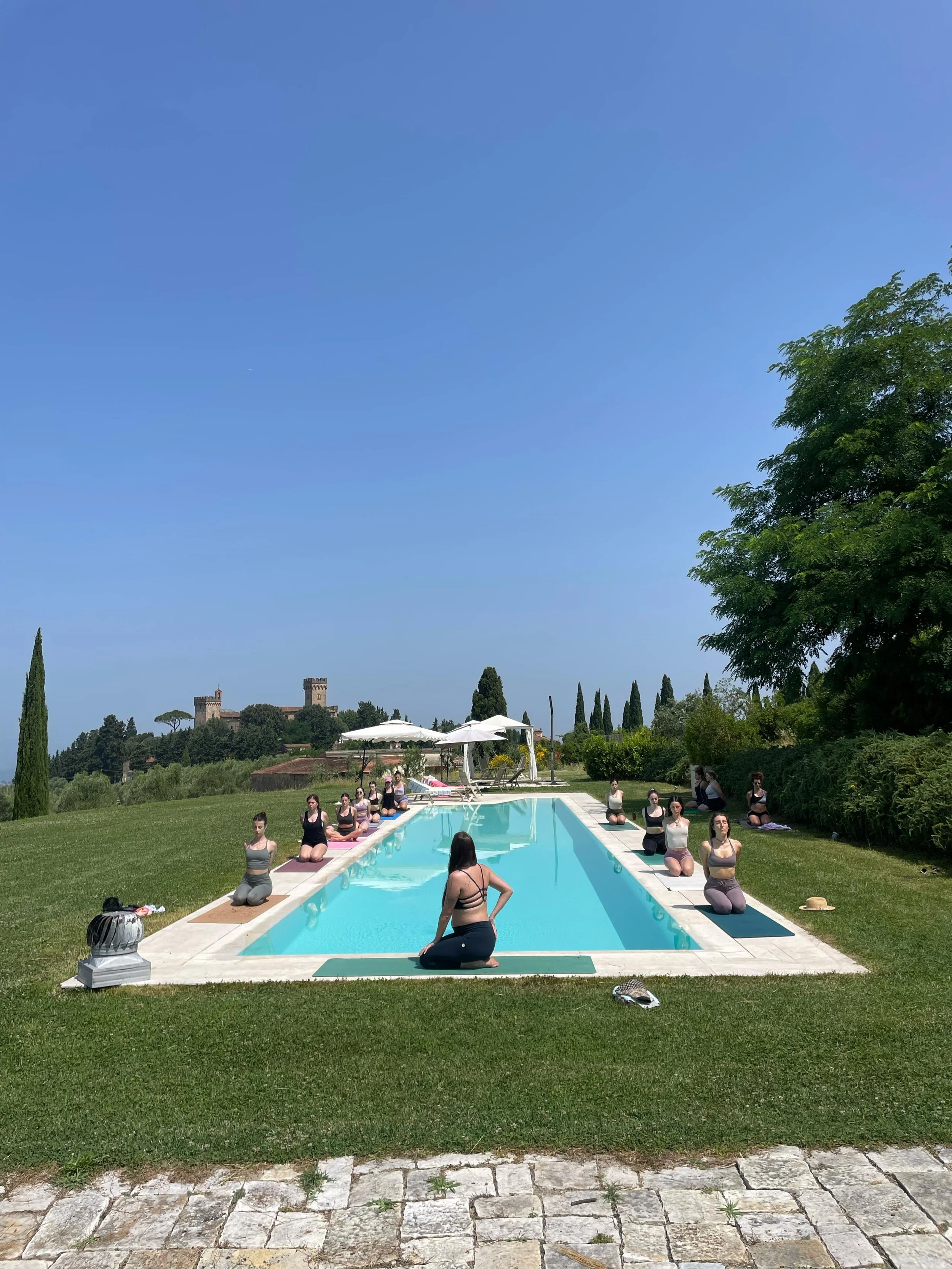 Persone che praticano yoga vicino a una piscina all'aperto in un giardino con alberi e un castello in collina sullo sfondo, in una giornata di sole.