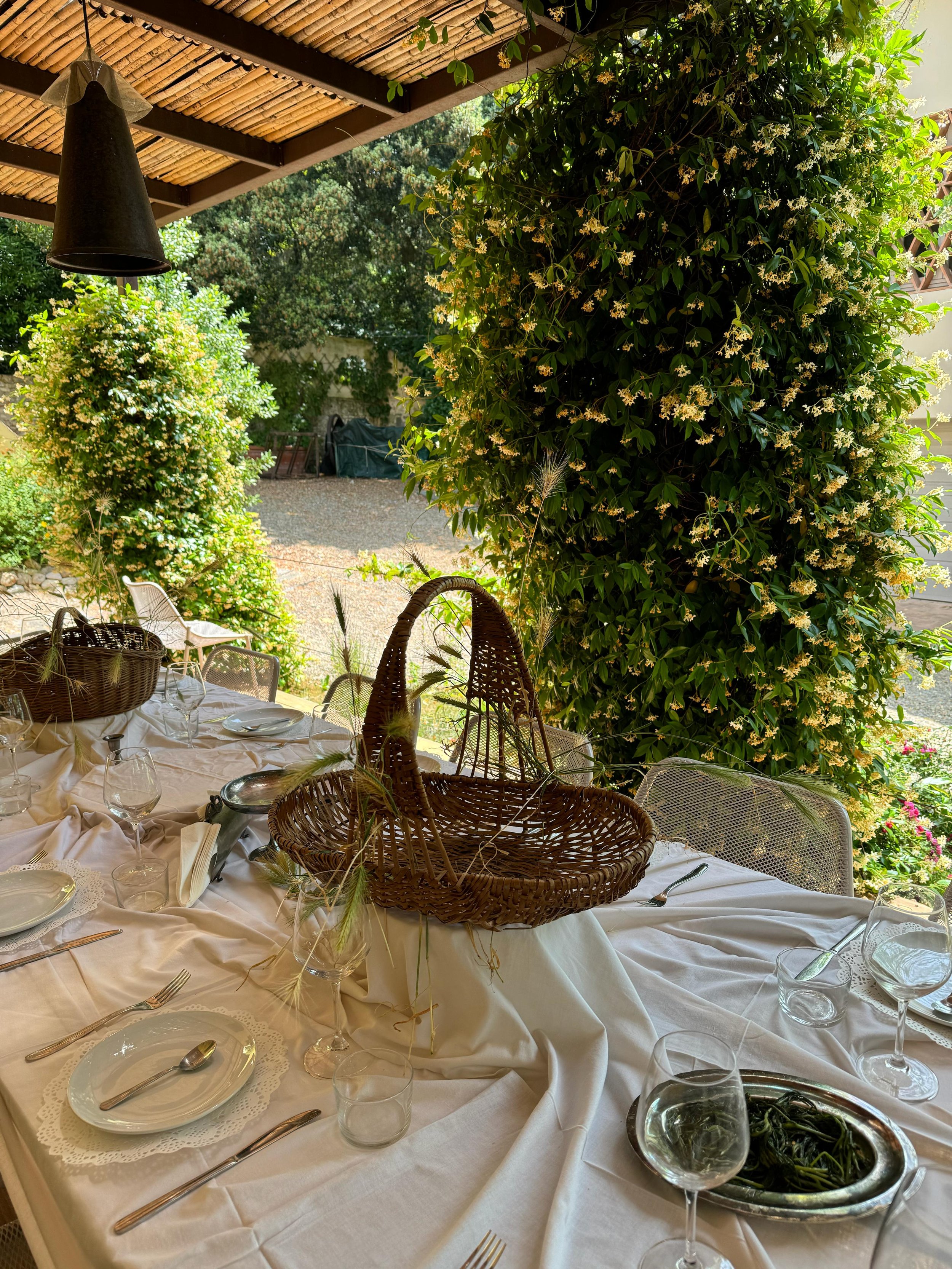Tavola imbandita all'aperto con stoviglie, bicchieri, posate, un cestino di vimini, in un ambiente naturale con vegetazione e piante in fiore.