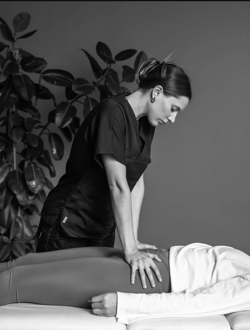 Una donna che esegue massaggi su una persona distesa, in un ambiente interno con piante sullo sfondo, in bianco e nero.
