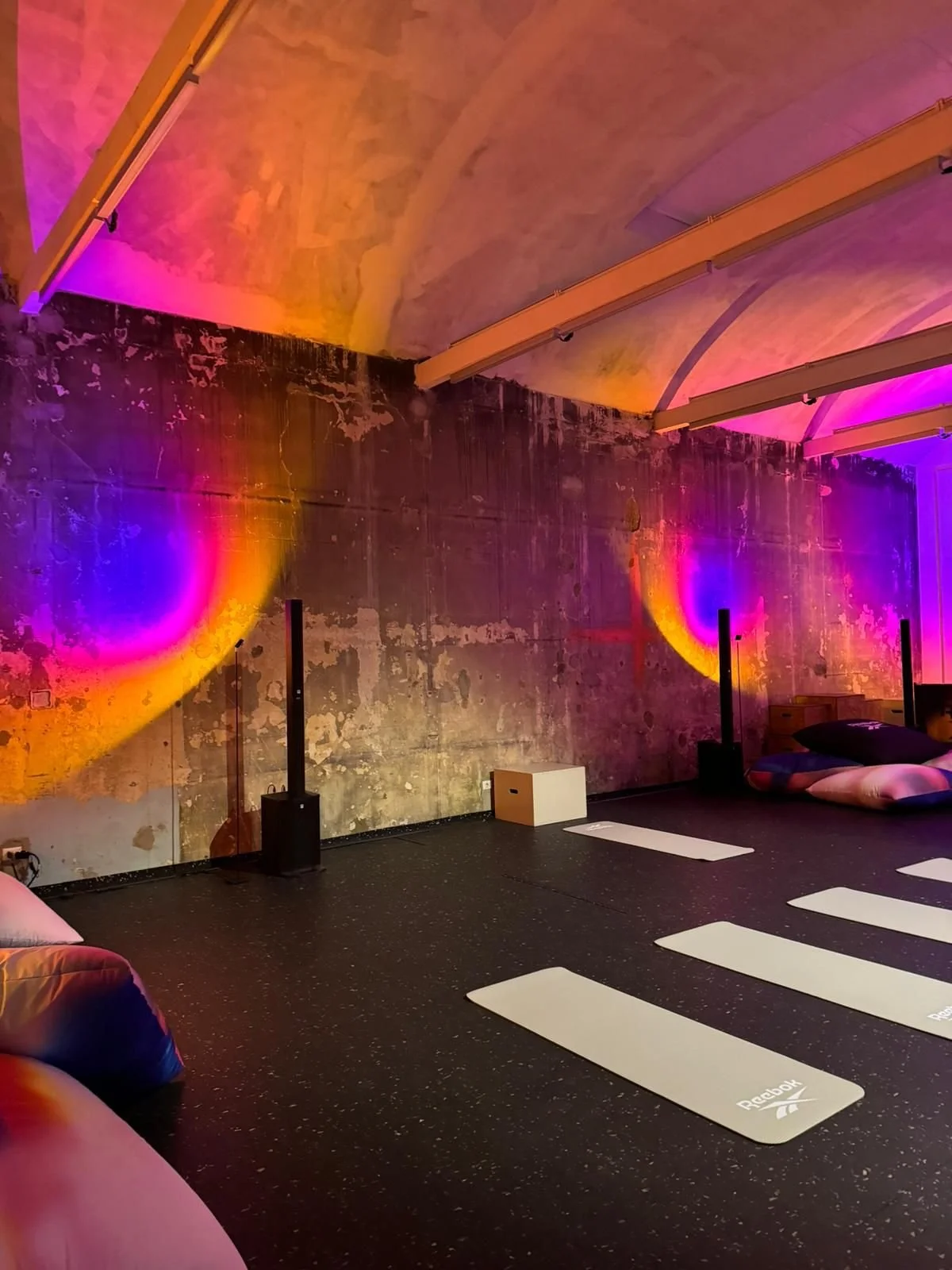 Interno di una sala per yoga con luci colorate e pareti in pietra. Ci sono quattro tappetini da yoga con il logo Reebok e cuscini ai bordi della stanza.