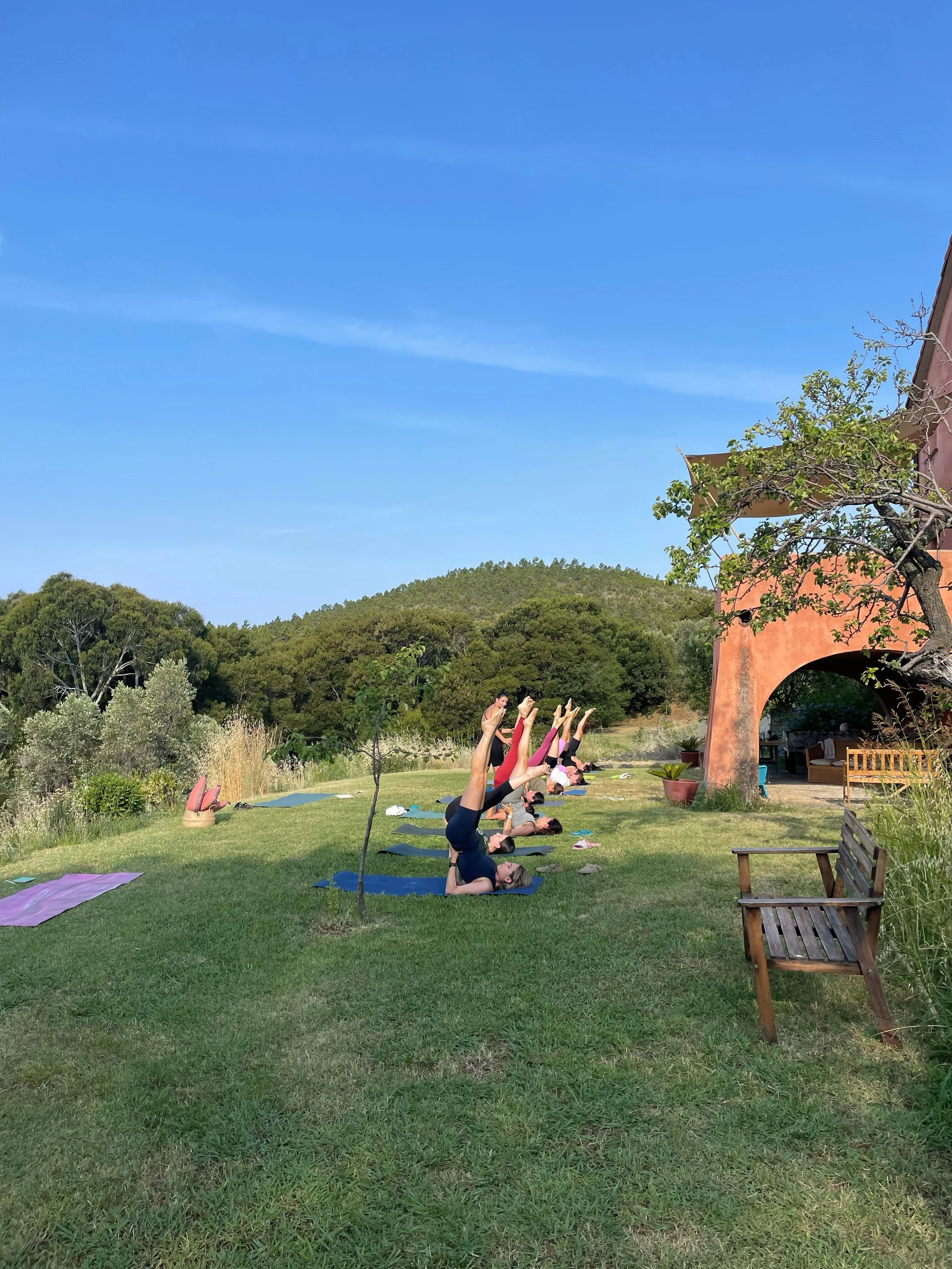 Persone che praticano yoga all'aperto su erba verde, con un cielo blu e colline sullo sfondo, vicino a una casa in stile mediterraneo con un'area esterna e piante.