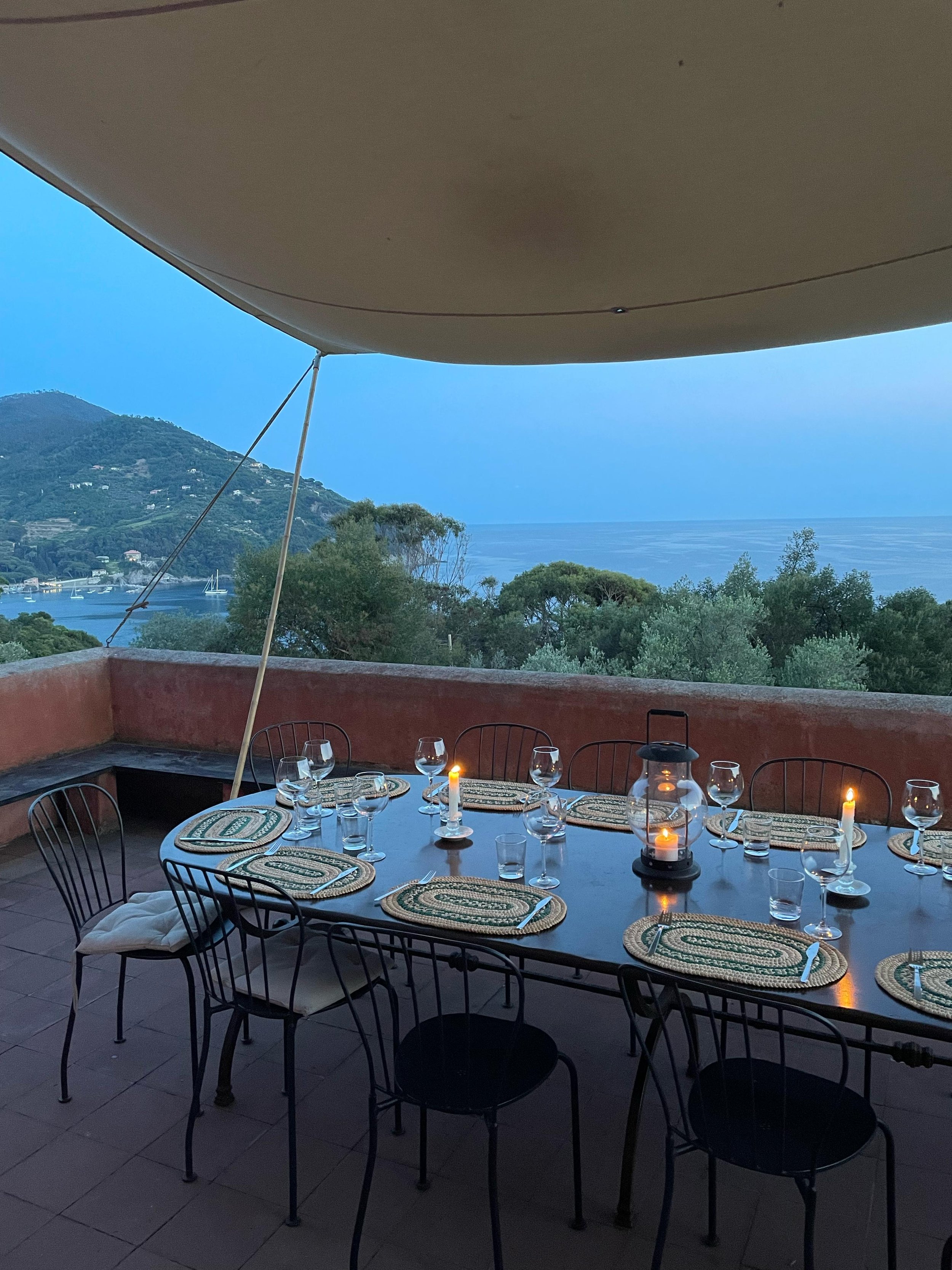 Tavolo da pranzo all'aperto con 10 posti, tovagliette, bicchieri e candele accese, con vista panoramica sul mare e sulle colline circostanti al tramonto.
