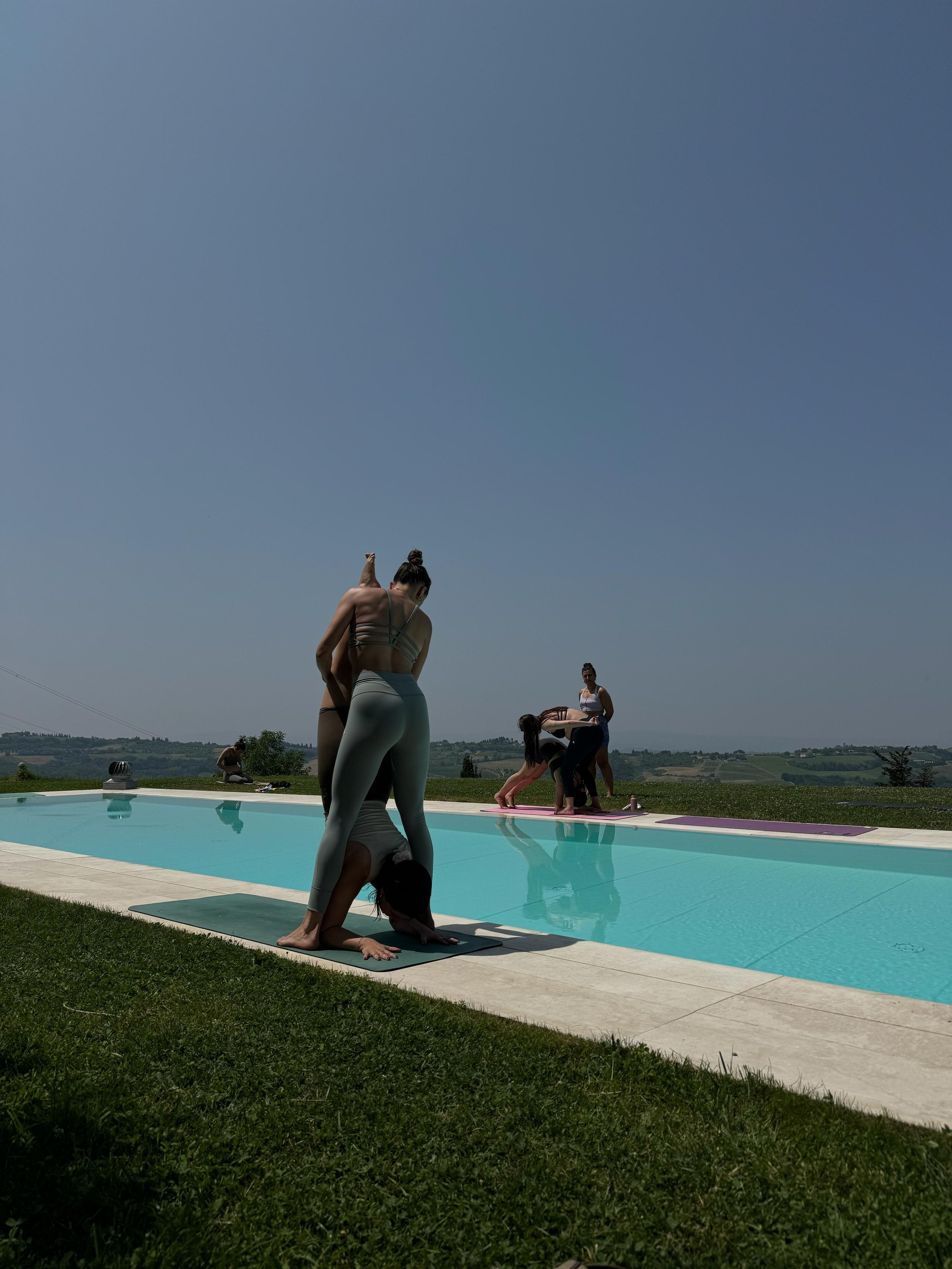 Persone che praticano yoga in una piscina all'aperto durante il giorno, con un paesaggio rurale sullo sfondo.
