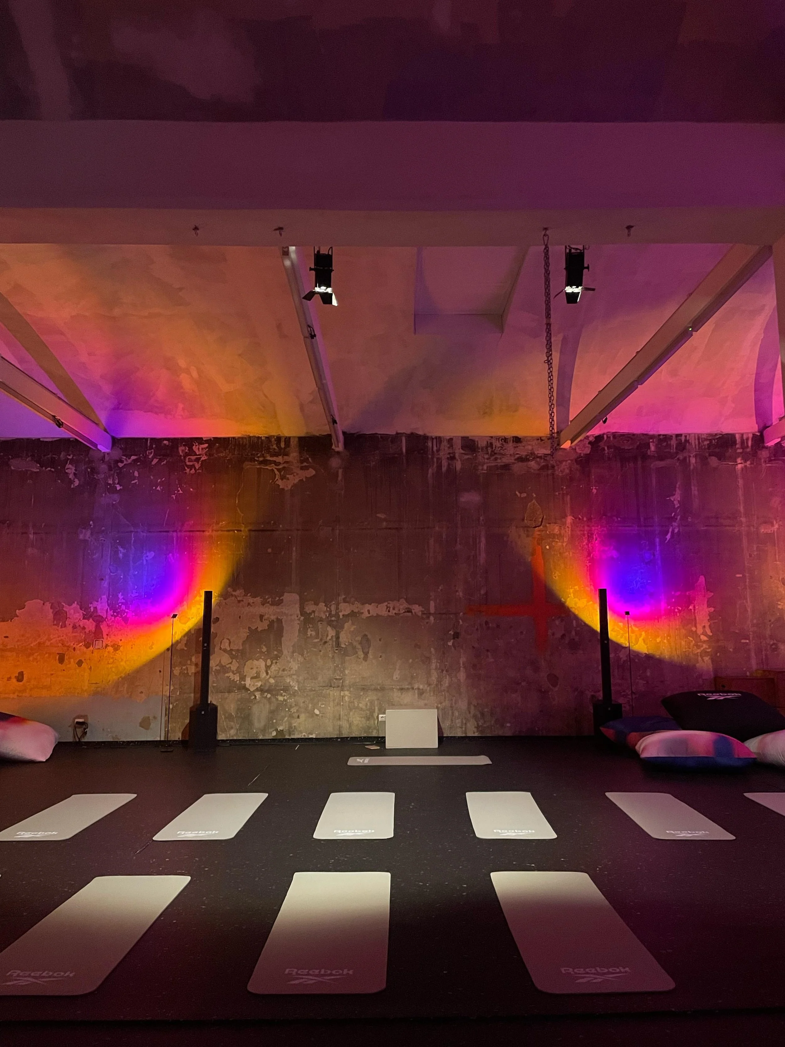 Stanza di yoga con tappetini Reebok disposti sul pavimento, luci colorate e parete in mattoni, con attrezzature per il fitness appese al soffitto.