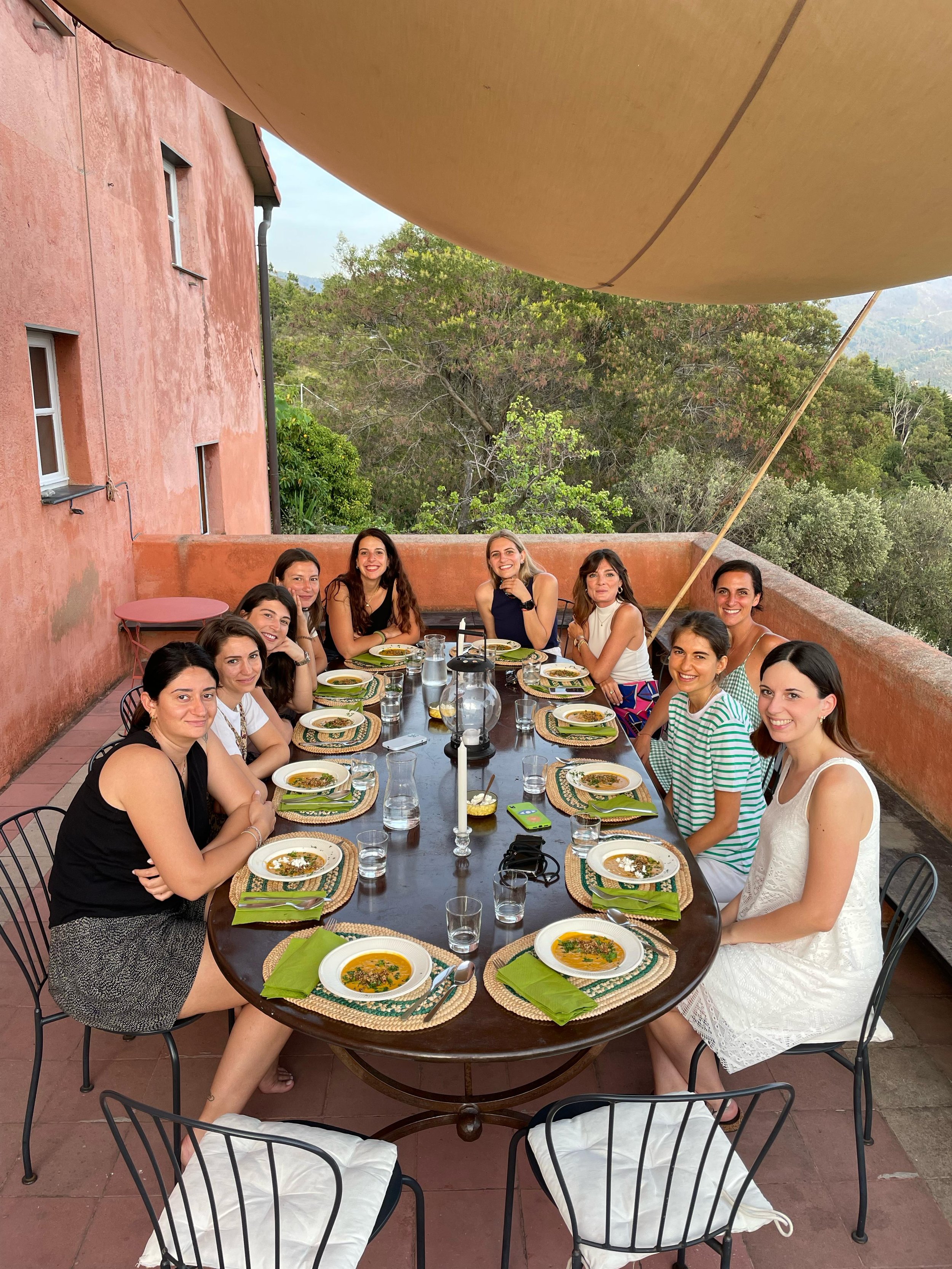 Gruppo di donne sedute attorno a un tavolo con zuppe, mentre mangiano e sorridono, in un terrazzo con vista naturale sullo sfondo