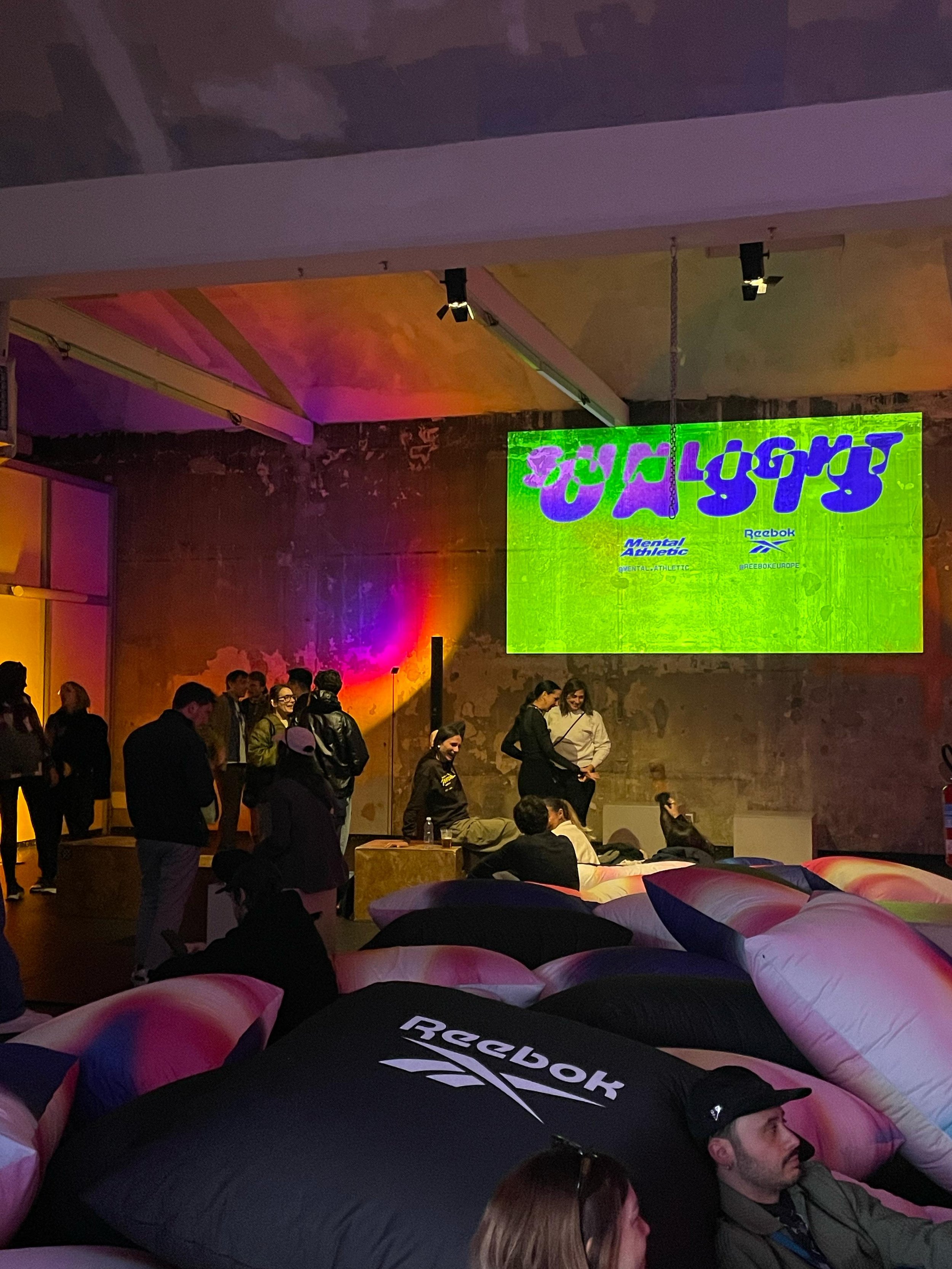 Ingresso evento con schermi luminosi e persone sedute e in piedi in un ambiente industriale con pareti di cemento, decorato con numerosi cuscini giganti in vari colori, con luci ambientali colorate.