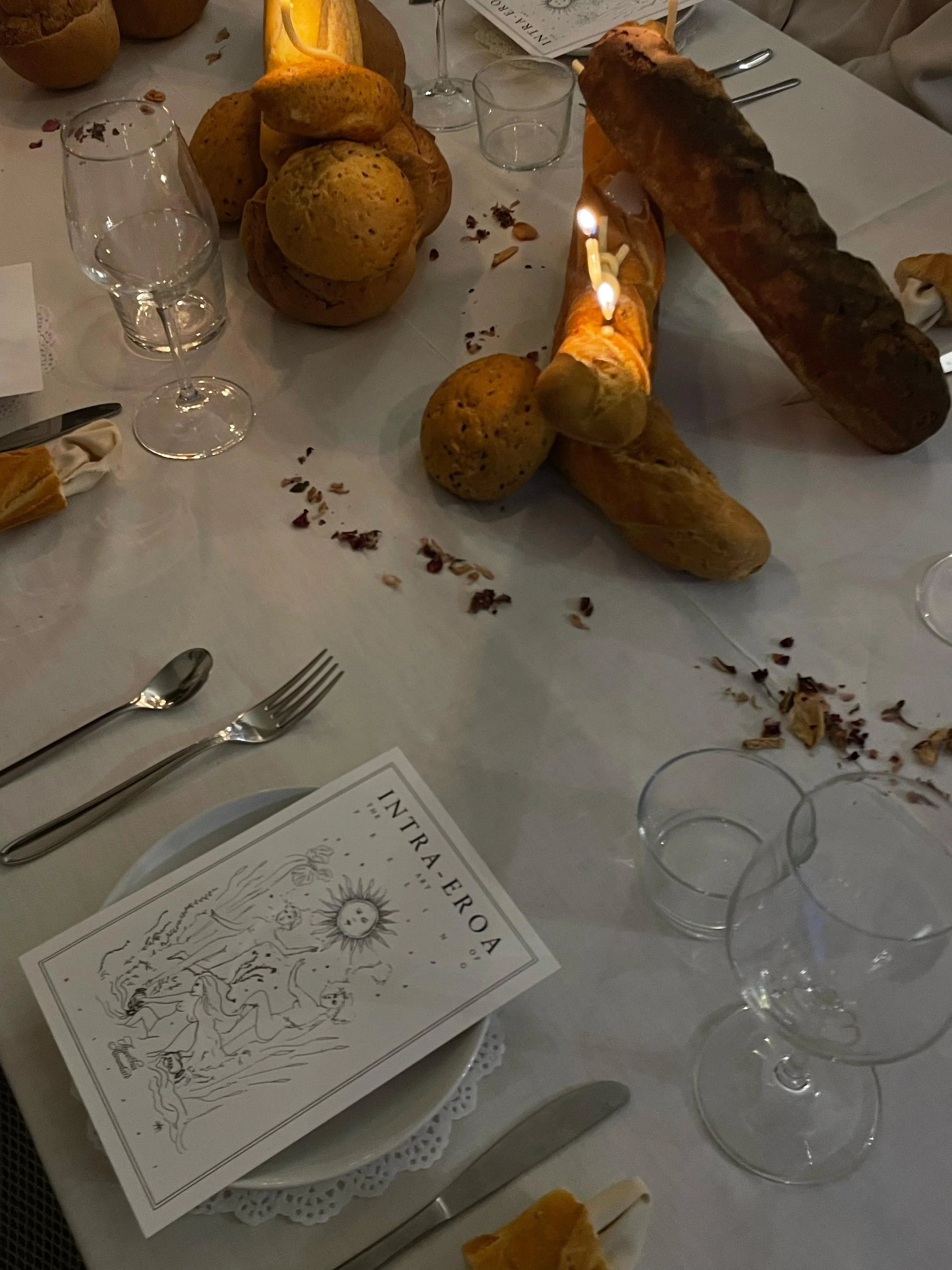 Tavola imbandita con pane, bicchieri vuoti e un menu con disegni, decorazioni floreali e un'illustrazione centrale, in un ambiente elegante.