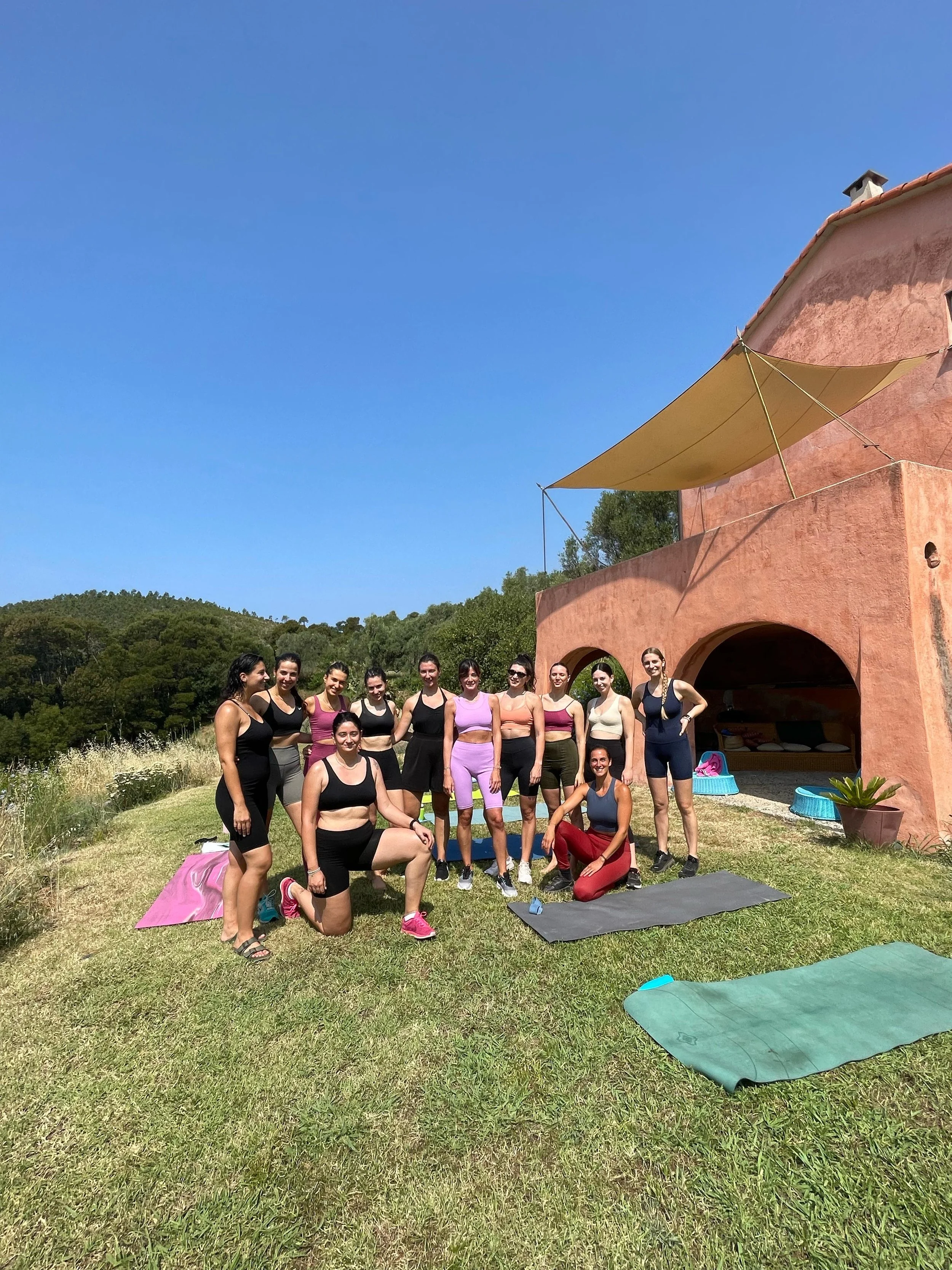 Un gruppo di donne in abbigliamento sportivo posano insieme all'aperto davanti a una casa di pietra rosa, con alcune stuoie da yoga sul prato verde, sotto un cielo sereno e azzurro.