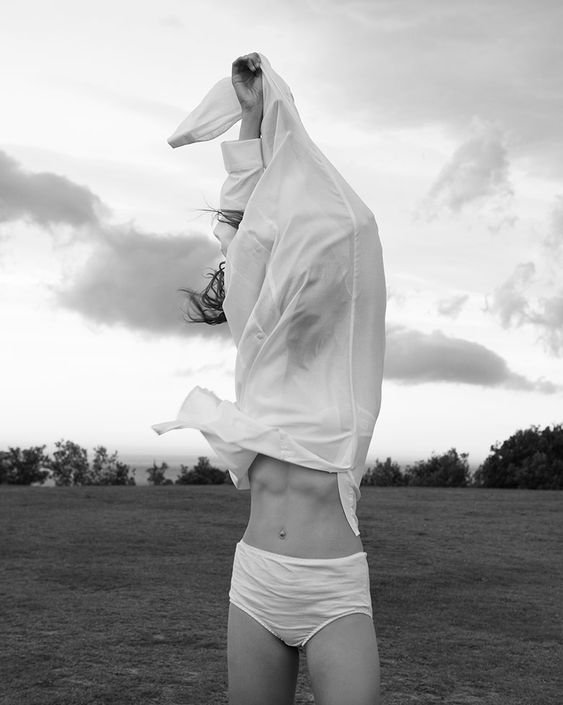 Donna in shorts con camicia bianca sollevata, in un campo aperto con nuvole nel cielo
