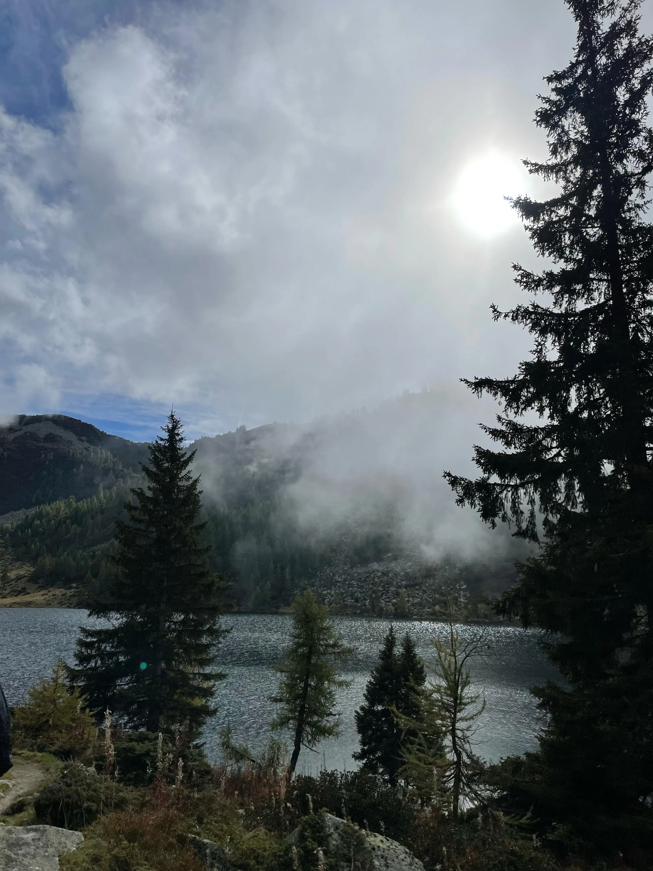 L'immagine mostra un paesaggio naturale con un lago circondato da alberi di conifere, montagne sullo sfondo e un cielo nuvoloso con il sole che filtra attraverso le nuvole.